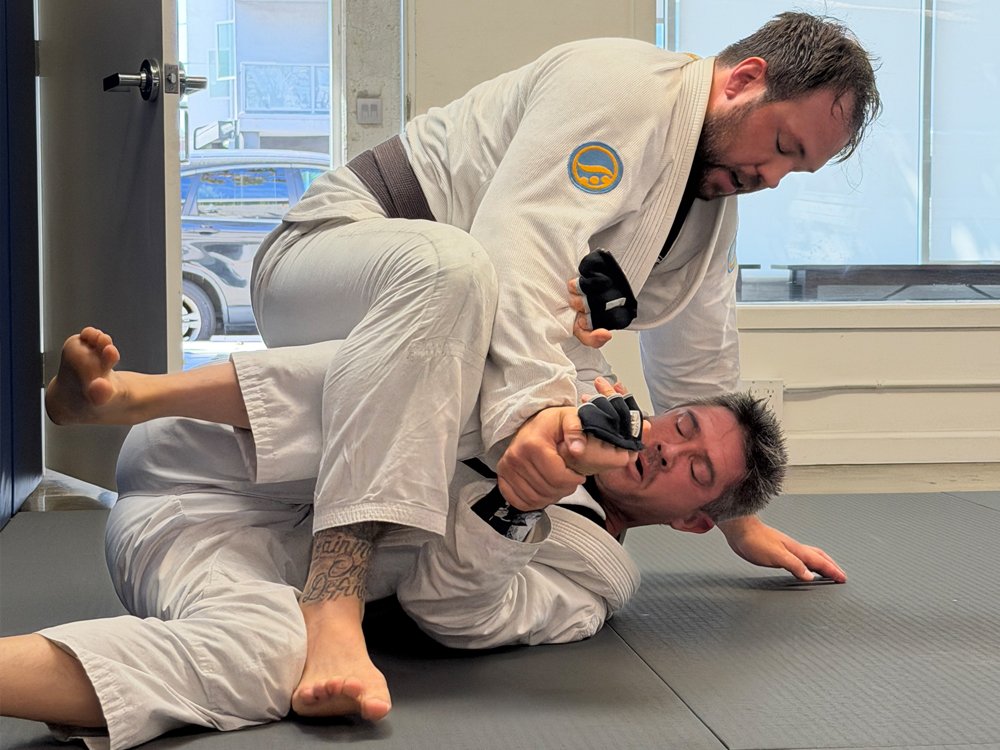 Two men practicing Brazilian Jiu-Jitsu on a black mat inside a martial arts gym. One man in a white gi is on the ground, and the other man in a white gi is pinned on top and appears to be attempting a submission move.