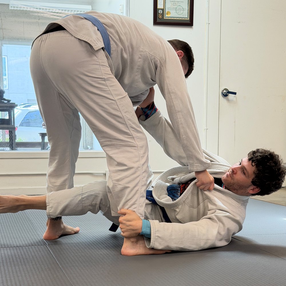 Two individuals practicing Brazilian Jiu-Jitsu in a martial arts gym, one person on the ground in a defensive position, and the other person attempting a submission hold.