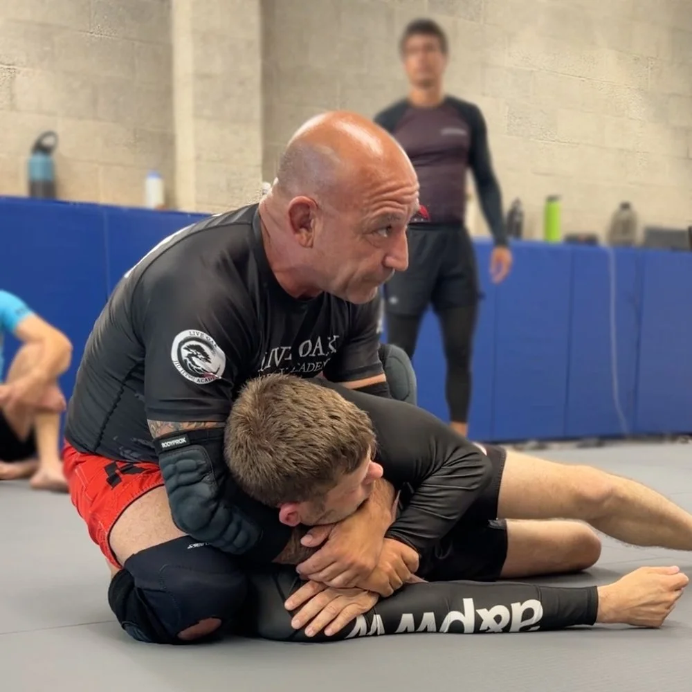 Two men practicing Brazilian Jiu-Jitsu on the mat, with one man on top and the other on the bottom, in a gym. A man in the background watches.