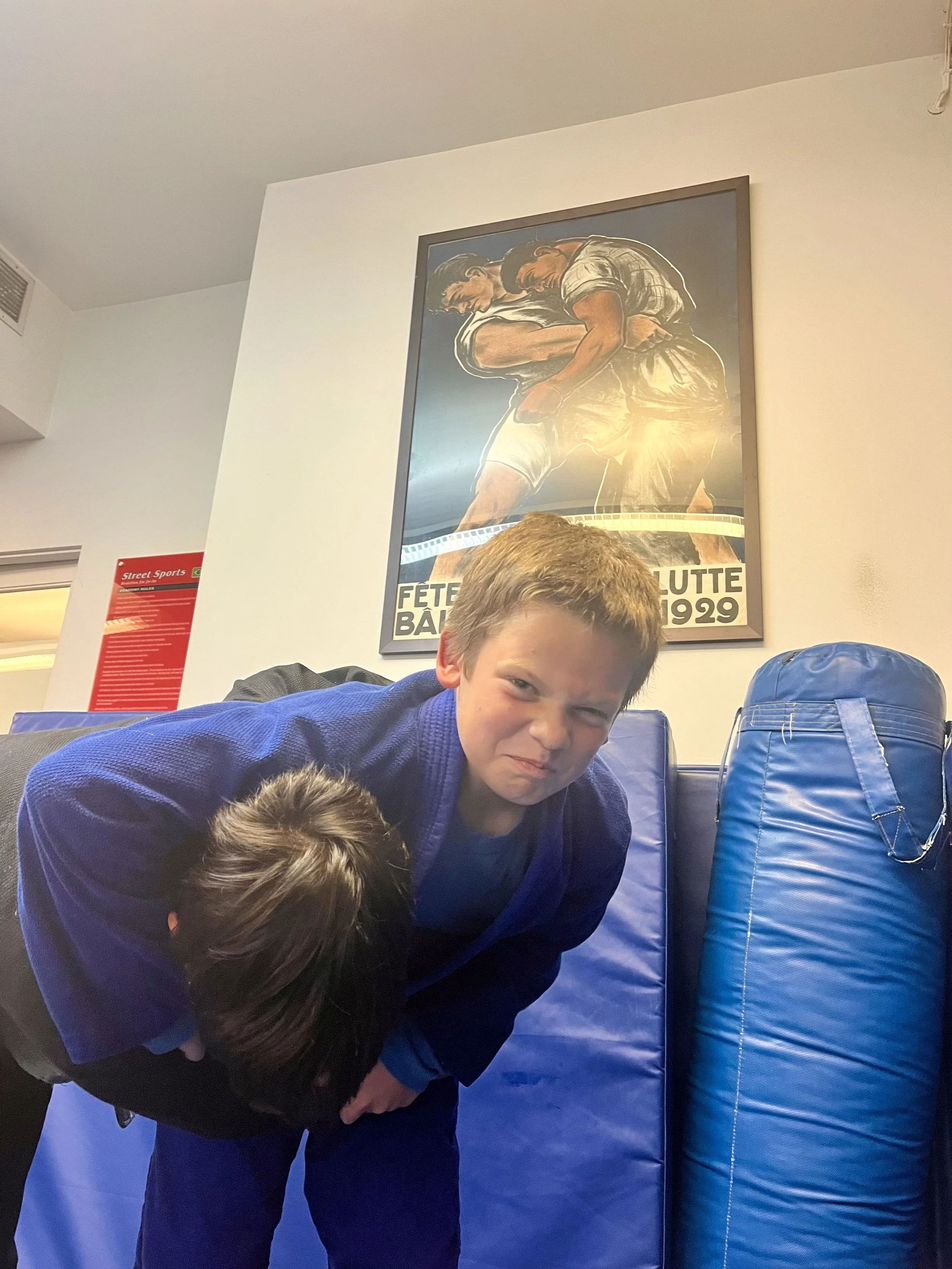 A boy in a blue gi practicing martial arts on blue padded mats in a gym, with another person leaning over him. A framed vintage poster of a wrestling match from 1929 hangs on the wall behind them.