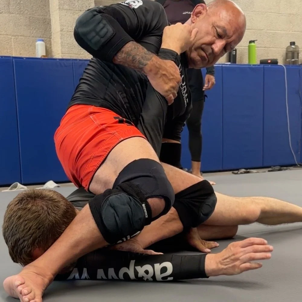 Two men practicing Brazilian Jiu-Jitsu on a gray mat in a gym. One man in black rash guard and red shorts is applying a choke to the other, who is on his stomach in a black gi with knee pads.