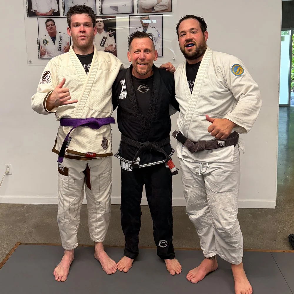 Three men in Brazilian Jiu-Jitsu gis standing on a mat, smiling, with the two taller men showing their thumbs up, one wearing a purple belt and the other a brown belt, in a martial arts gym.