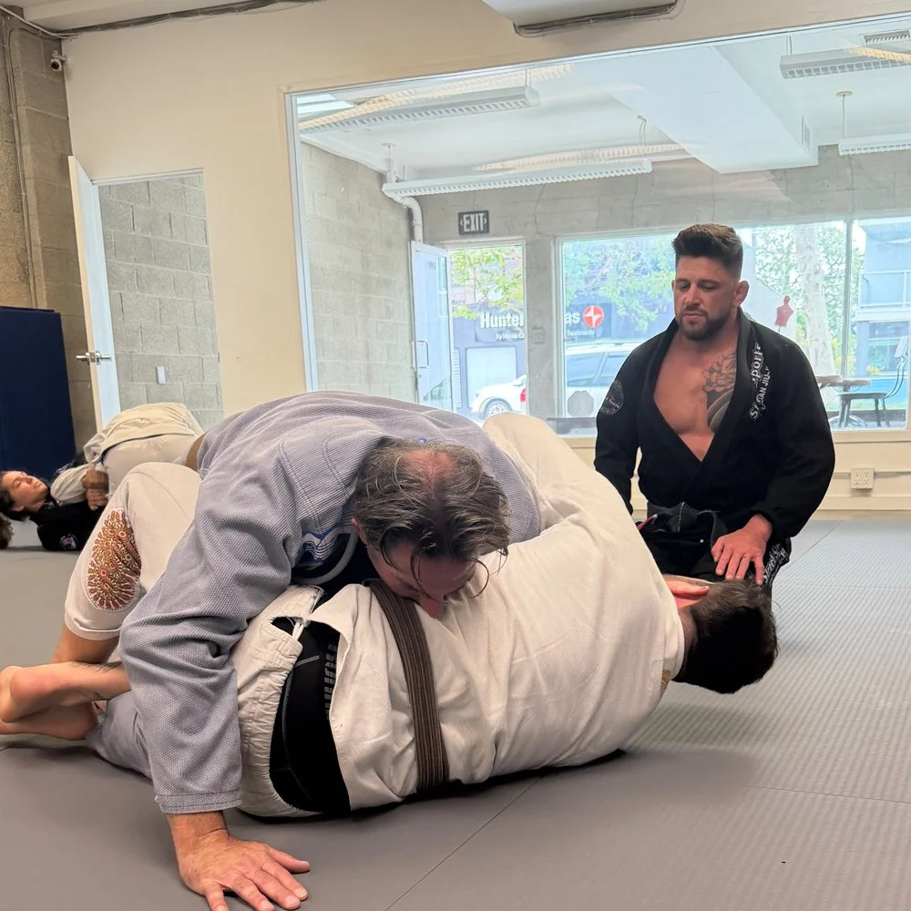 Two men practicing Brazilian Jiu-Jitsu on a mat during a class, with one man applying a choke and the other on the ground, while another person is lying on a nearby mat.
