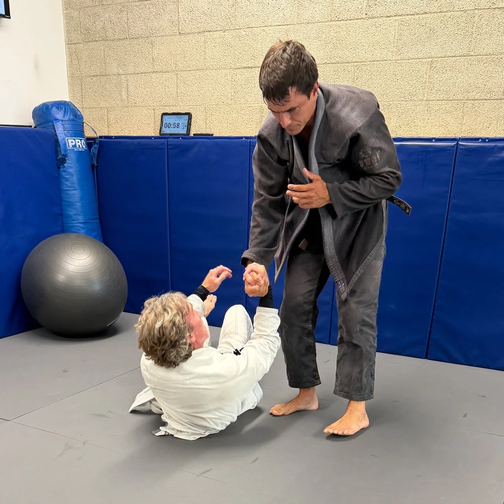 Two men practicing Brazilian Jiu-Jitsu in a training gym. One man in a white gi on the floor, and the other man in a black gi standing, holding his opponent's wrist.