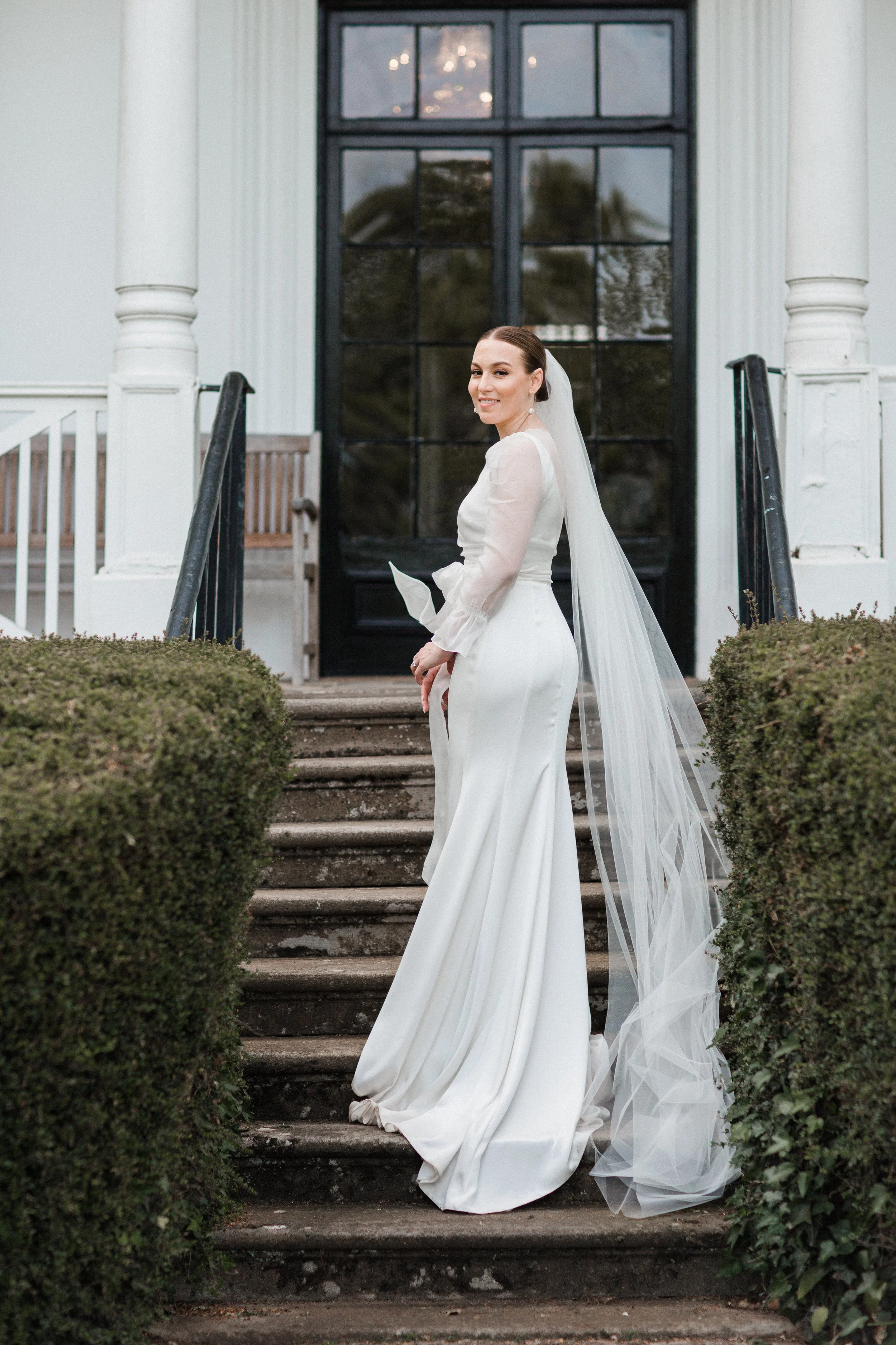 A bride in a white wedding dress with long sleeves and a veil standing on stairs in front of a large black door at a white building.