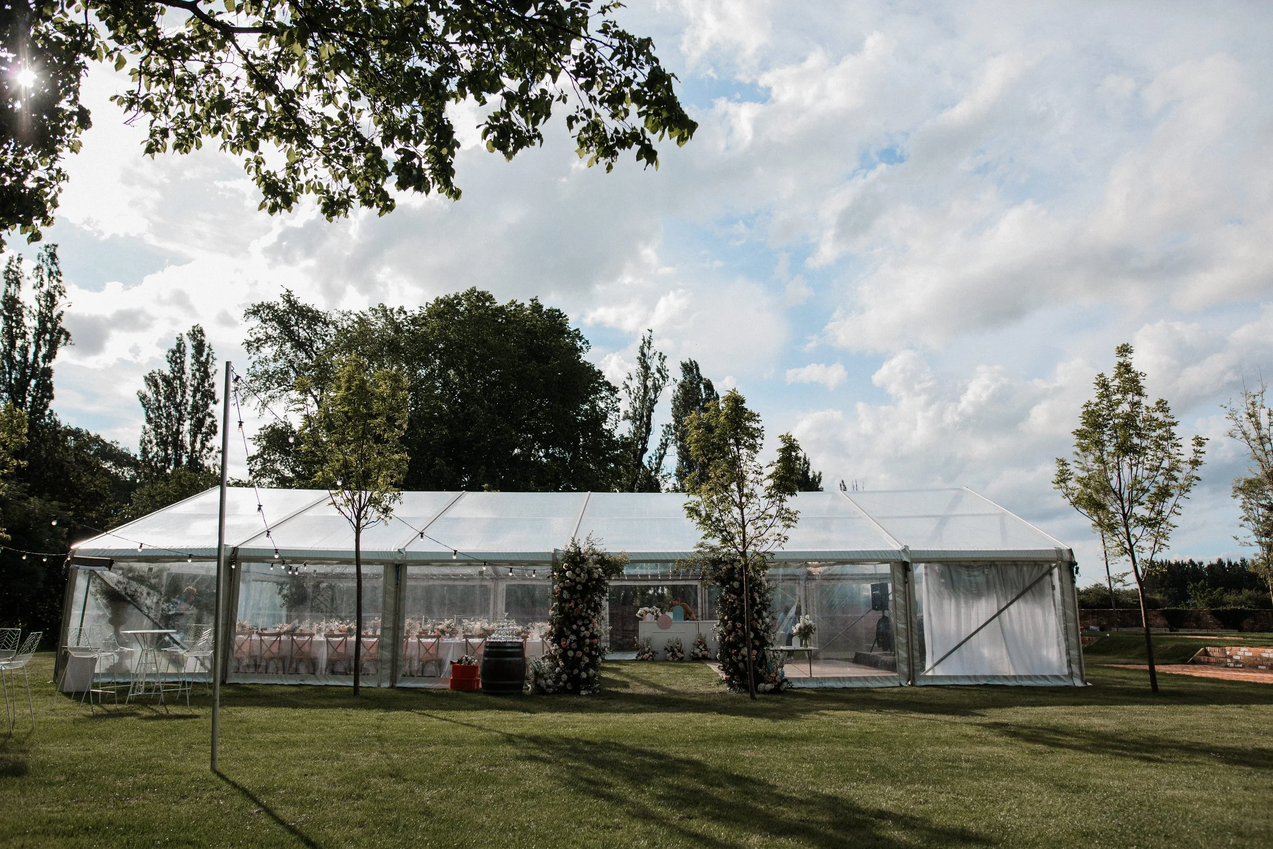 Outdoor event tent set up on a grassy field with trees and cloudy sky in the background, decorated with floral arrangements at the entrance.