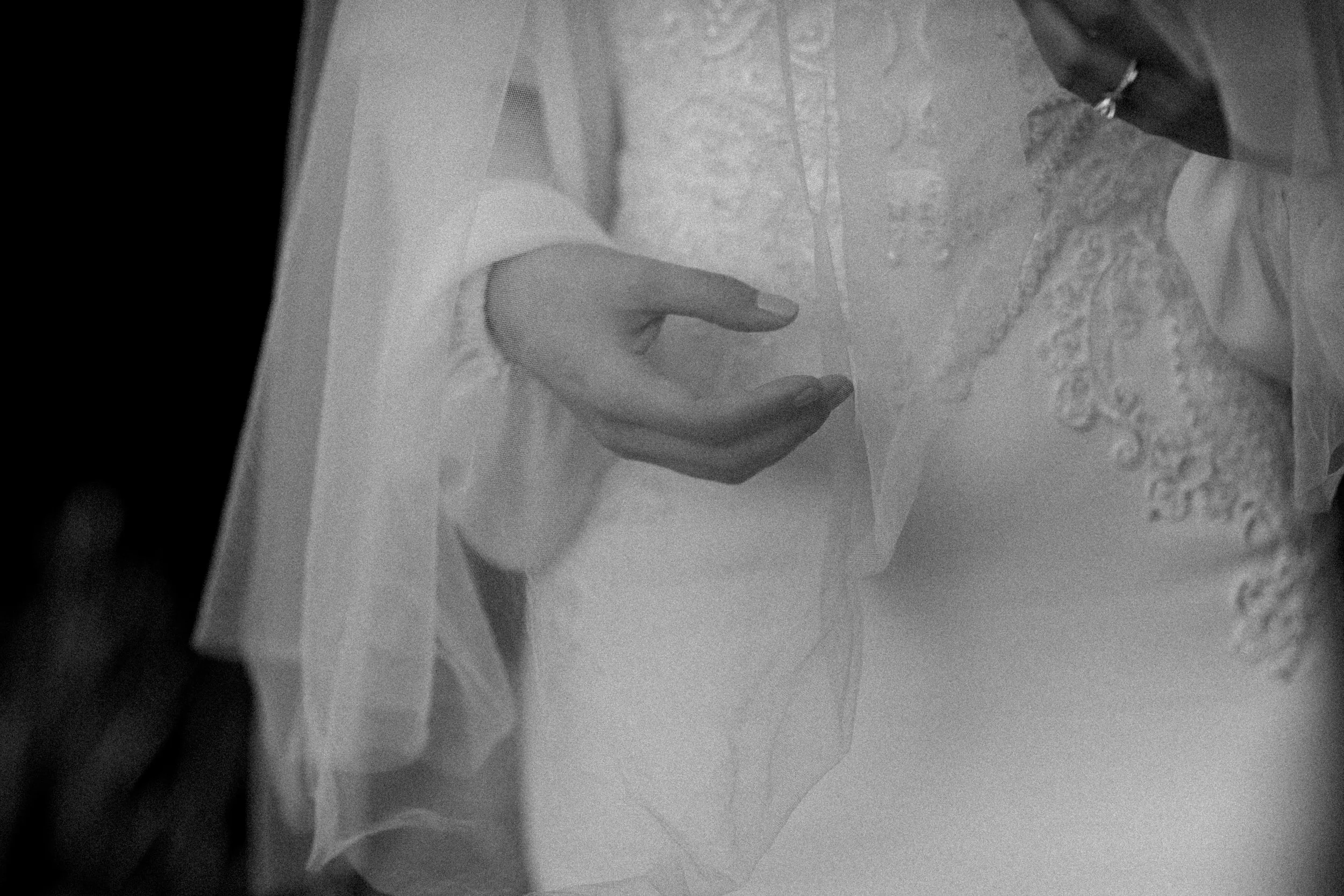 Close-up of a person's hand and part of their wedding dress, with lace details, fabric, and a ring visible on their finger.