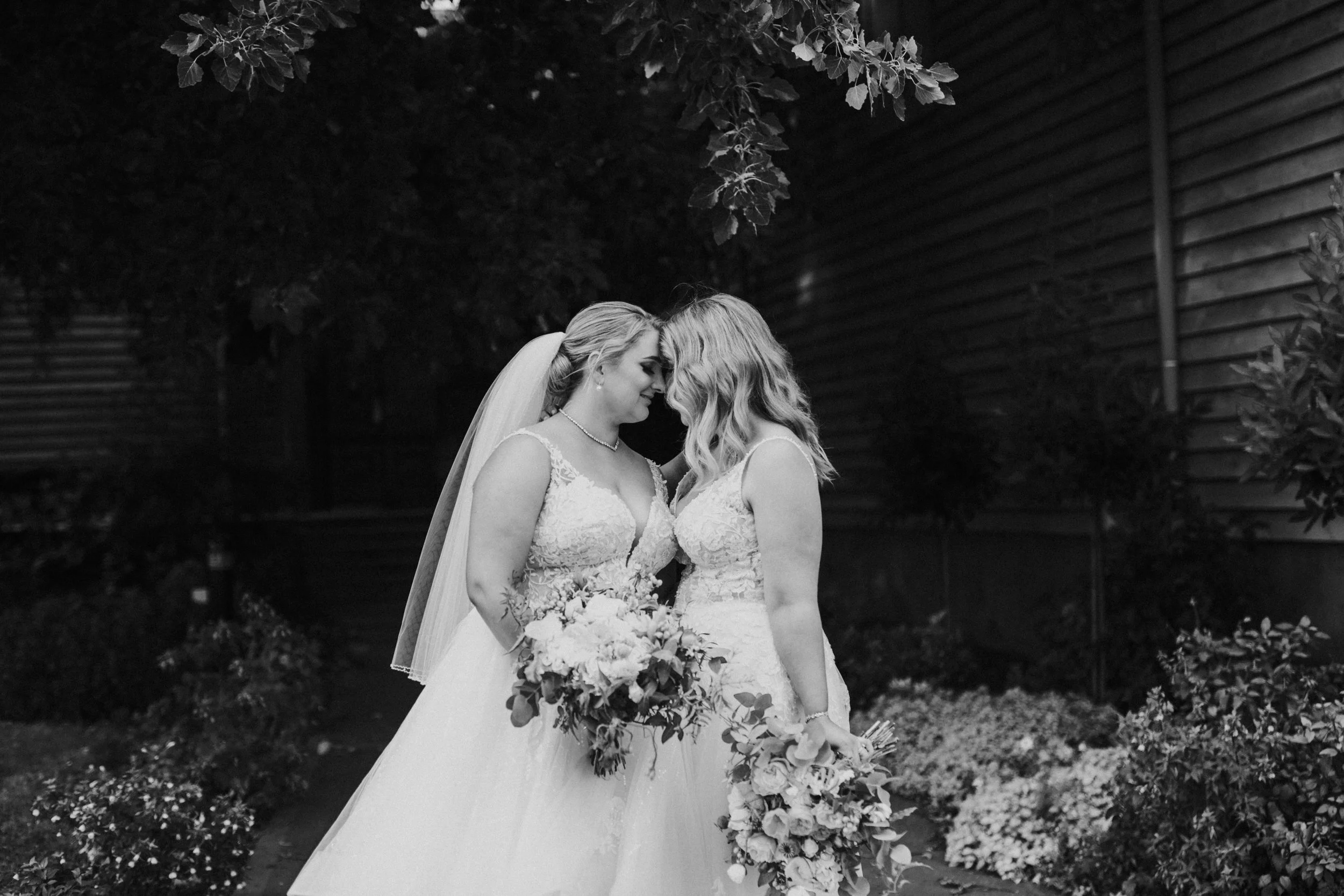 Two women in wedding dresses touching foreheads in a garden at night.