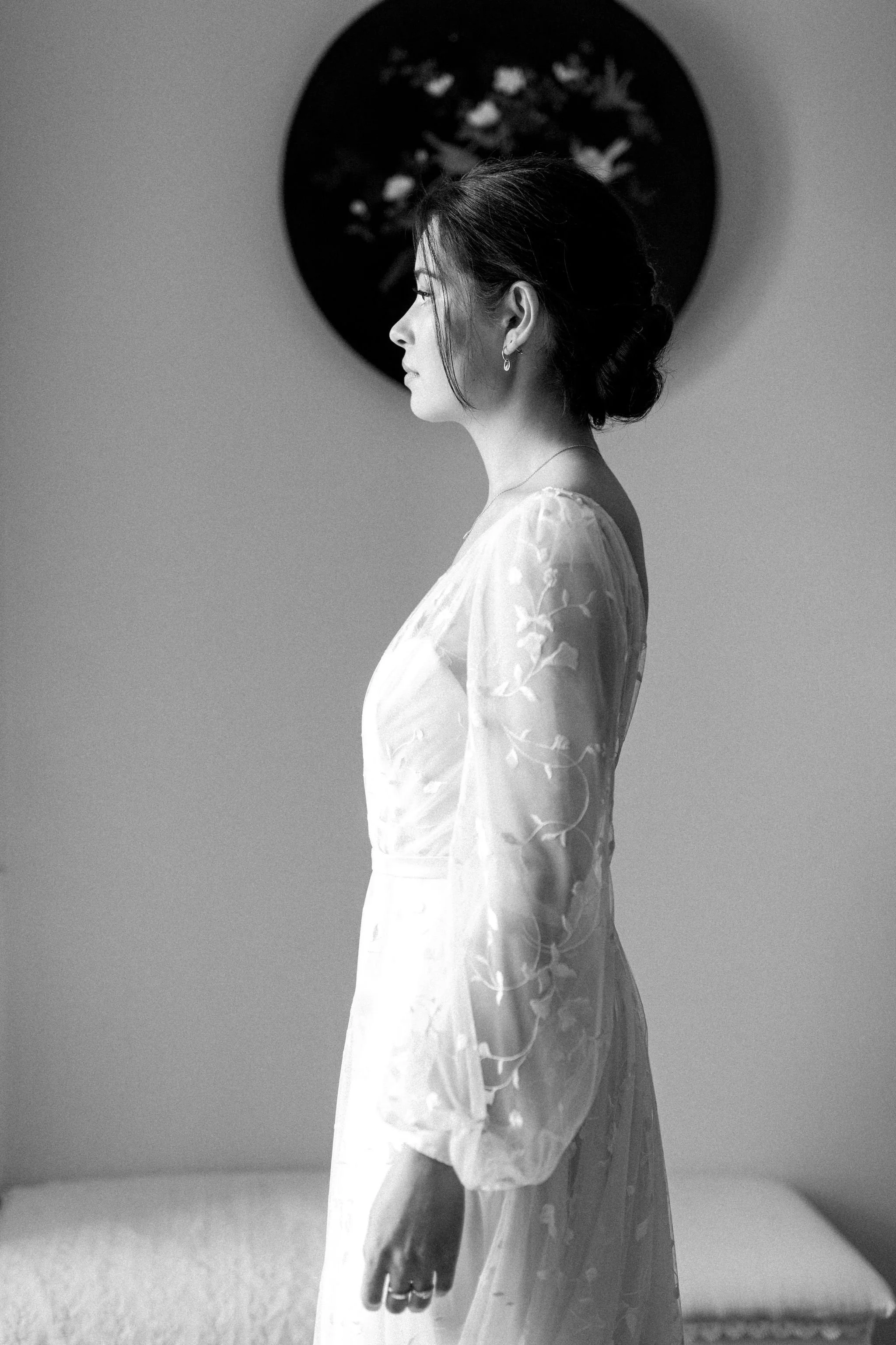 A black and white photo of a woman in a delicate, embroidered dress with long sheer sleeves, standing sideways in profile. She has dark hair styled in an elegant updo and wears earrings and a ring.