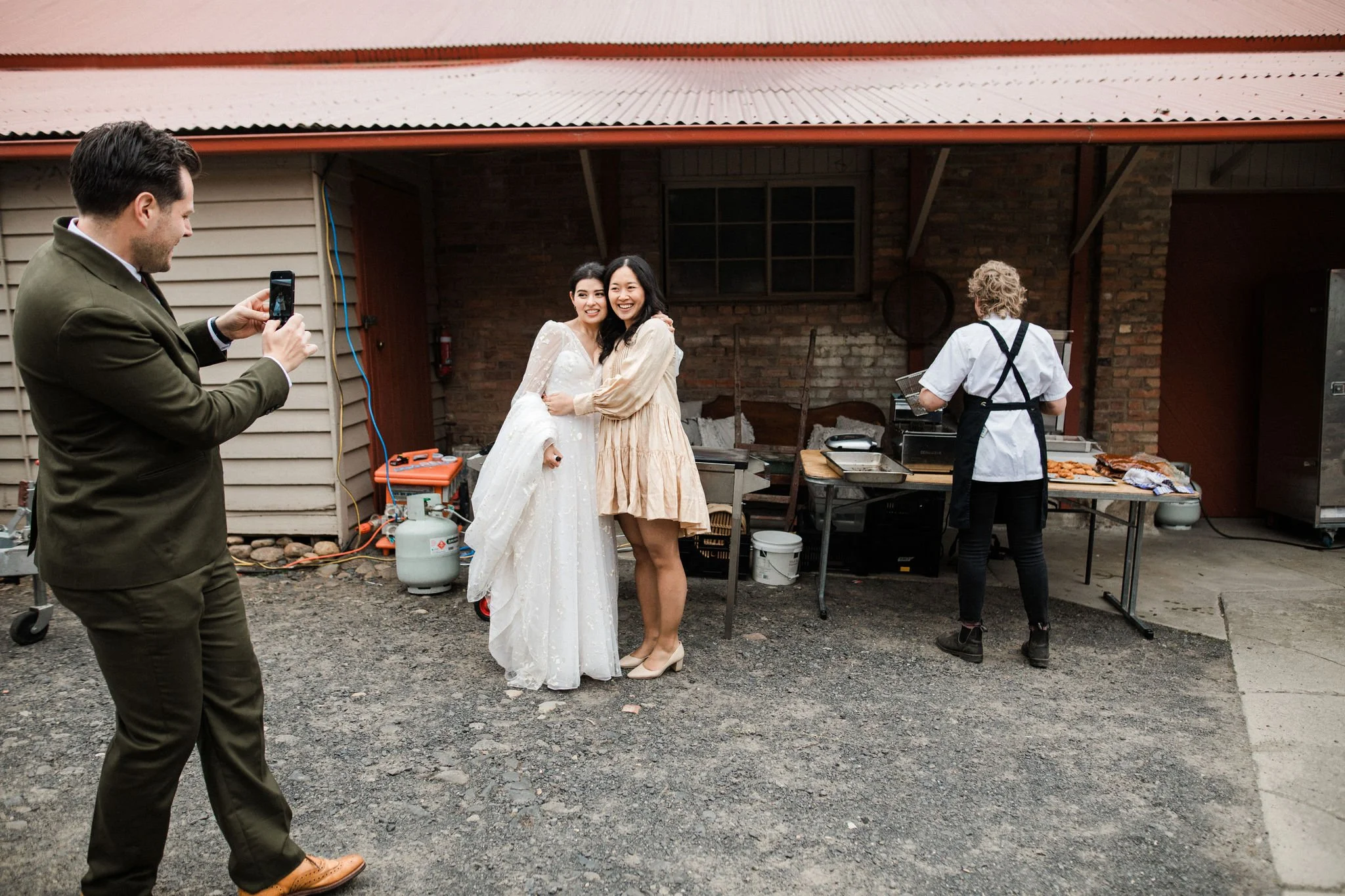 Two women, one in a wedding dress and the other in a beige dress, posing and smiling for a photo taken by a man in a green suit in an outdoor rustic setting. A person cooking at a grill is seen in the background.