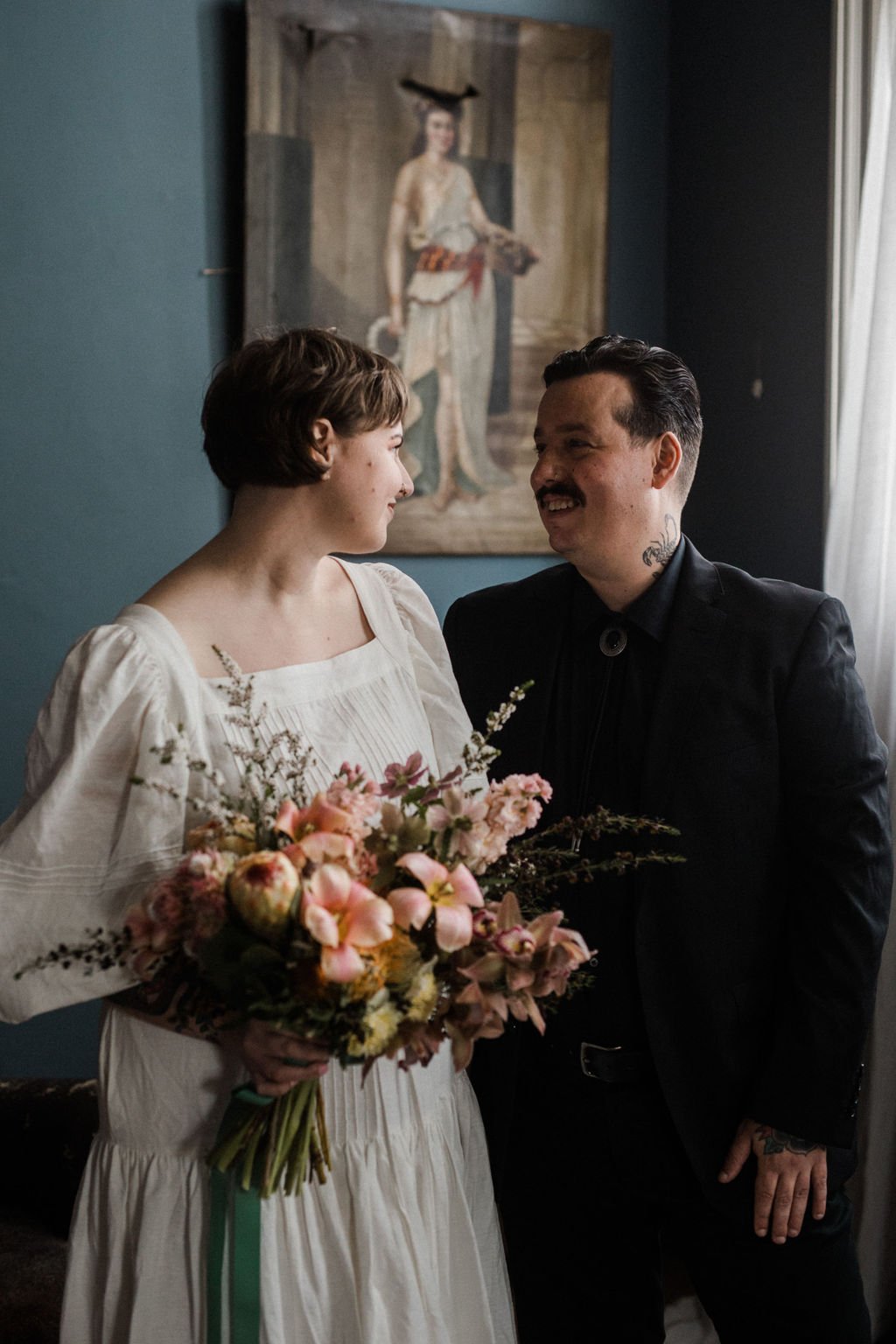 A couple dressed in vintage-style clothing shares a tender moment indoors, with the woman holding a bouquet of flowers and looking at the man who is smiling back at her, in front of a painting of a woman in historical attire.