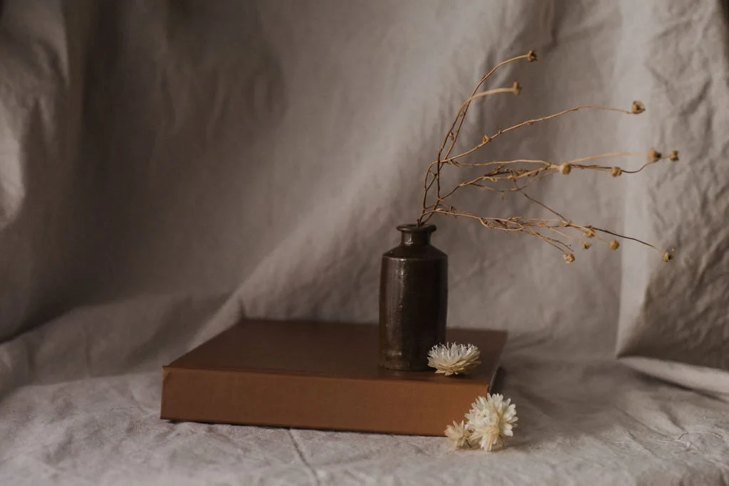 A dark brown vase with dry twigs sits on a brown rectangular box, with white flowers beside it, against a beige textured background.