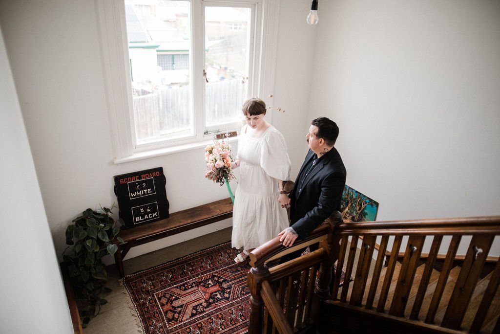 A woman in a white dress holding a bouquet of flowers walking down a staircase holding hands with a man in a dark suit inside a home.