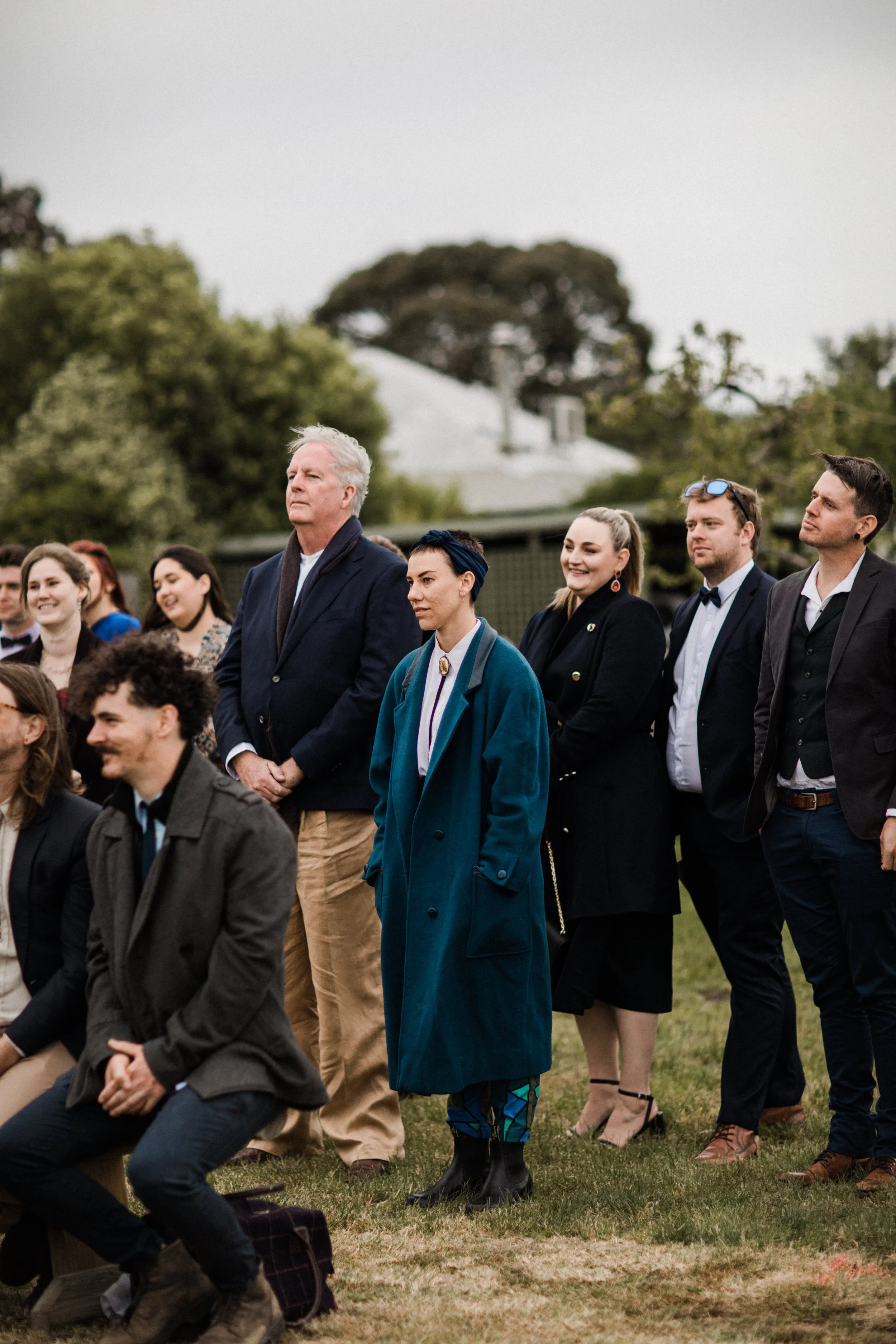 People attending an outdoor event, standing on grass, dressed in formal and semi-formal attire, with trees and a house in the background.