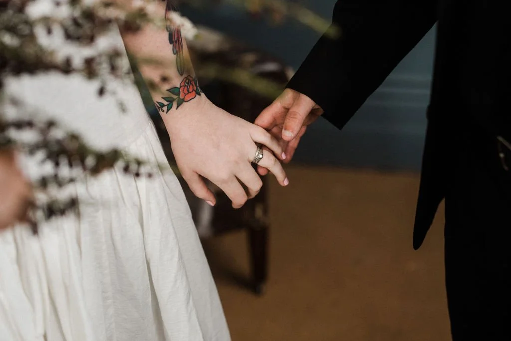 A couple holding hands, the woman wearing a wedding ring and with a tattooed arm, the man in a black coat, indoors.