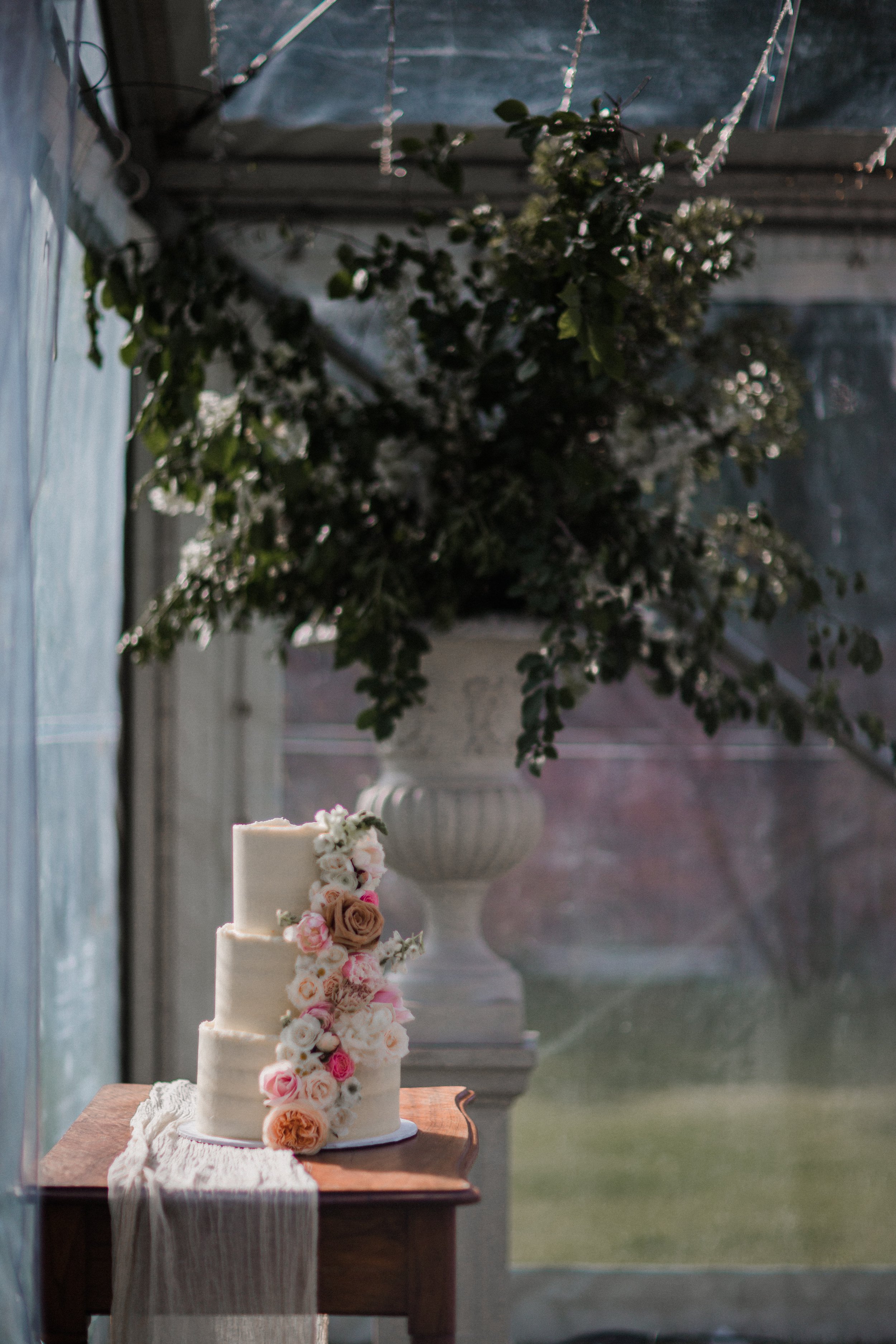 A three-tiered wedding cake decorated with pink, white, and peach roses, and a brown rose on top, placed on a wooden table with a white doily. In the background, there is a large outdoor urn with green foliage, set inside a glass structure with sunli