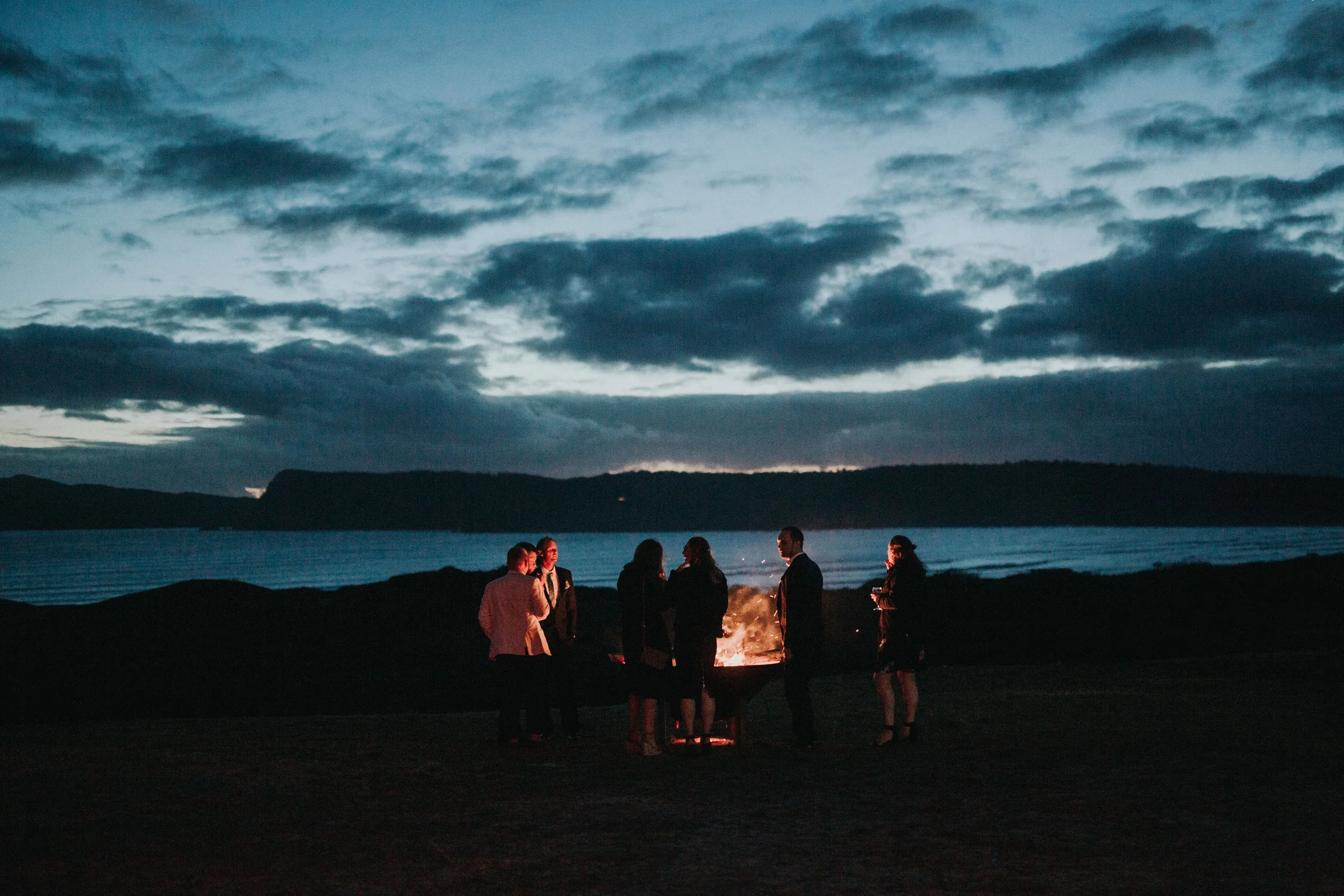 A group of people gathered around a campfire on a beach at dusk, with a dark sky and water in the background.