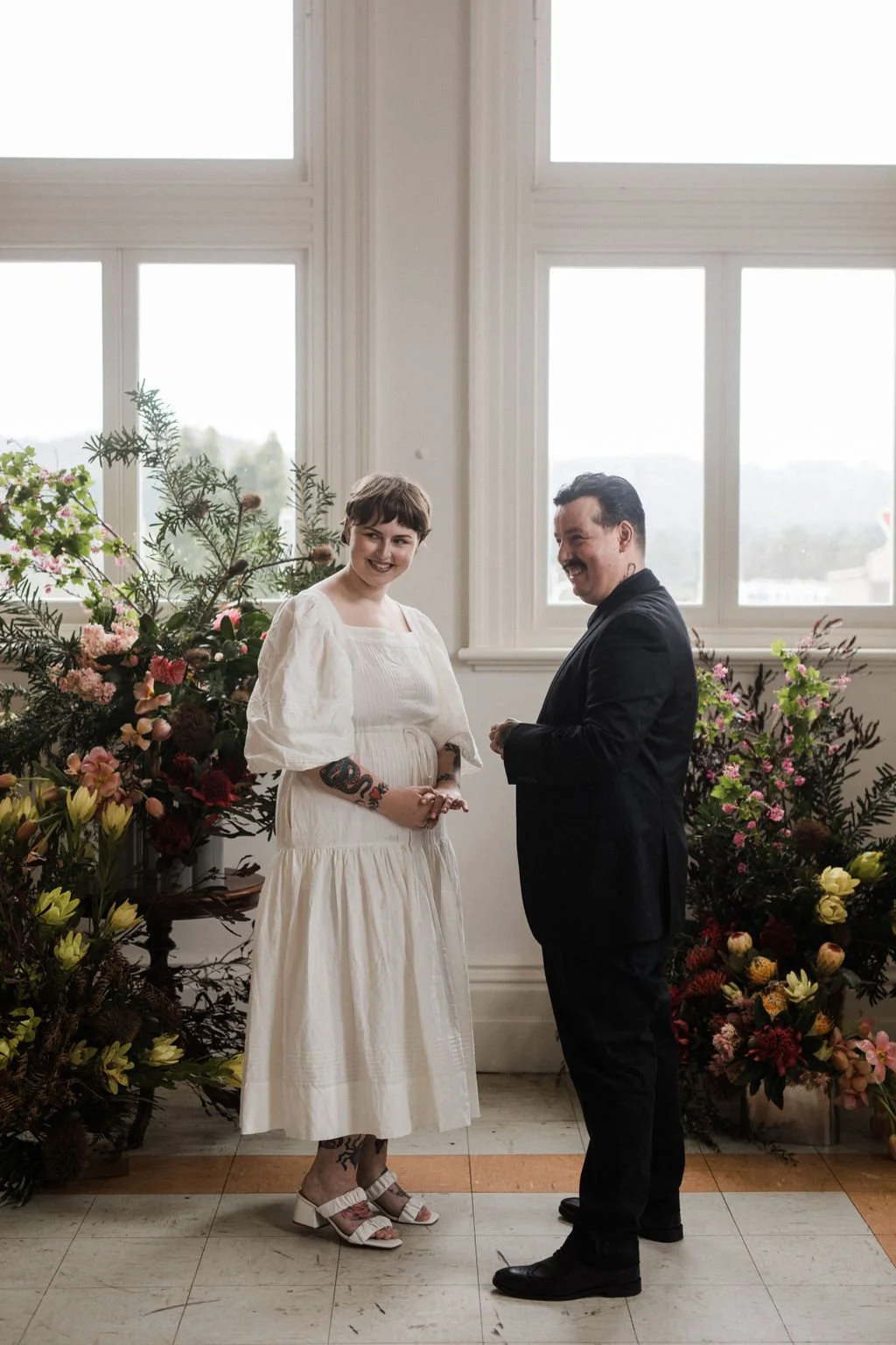 A couple standing indoors, surrounded by large floral arrangements, with large windows behind them. The woman wears a white dress with puffy sleeves and heels, and has tattoos on her arms and legs. The man wears a black suit and shoes, smiling at eac