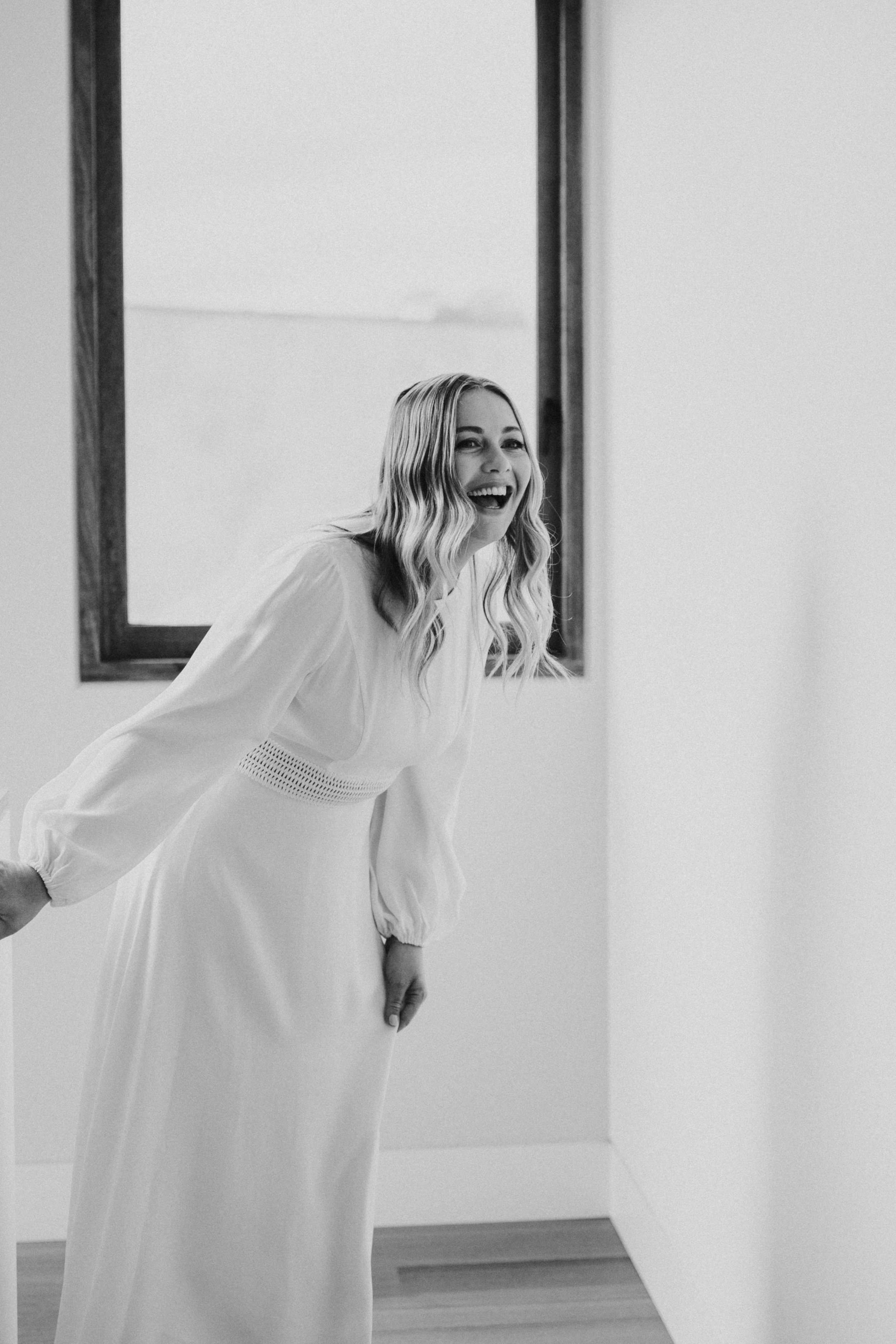 Black and white photo of a smiling woman in a long, flowing dress, leaning forward with her hands on her knees, standing in a room with plain walls and a window behind her.