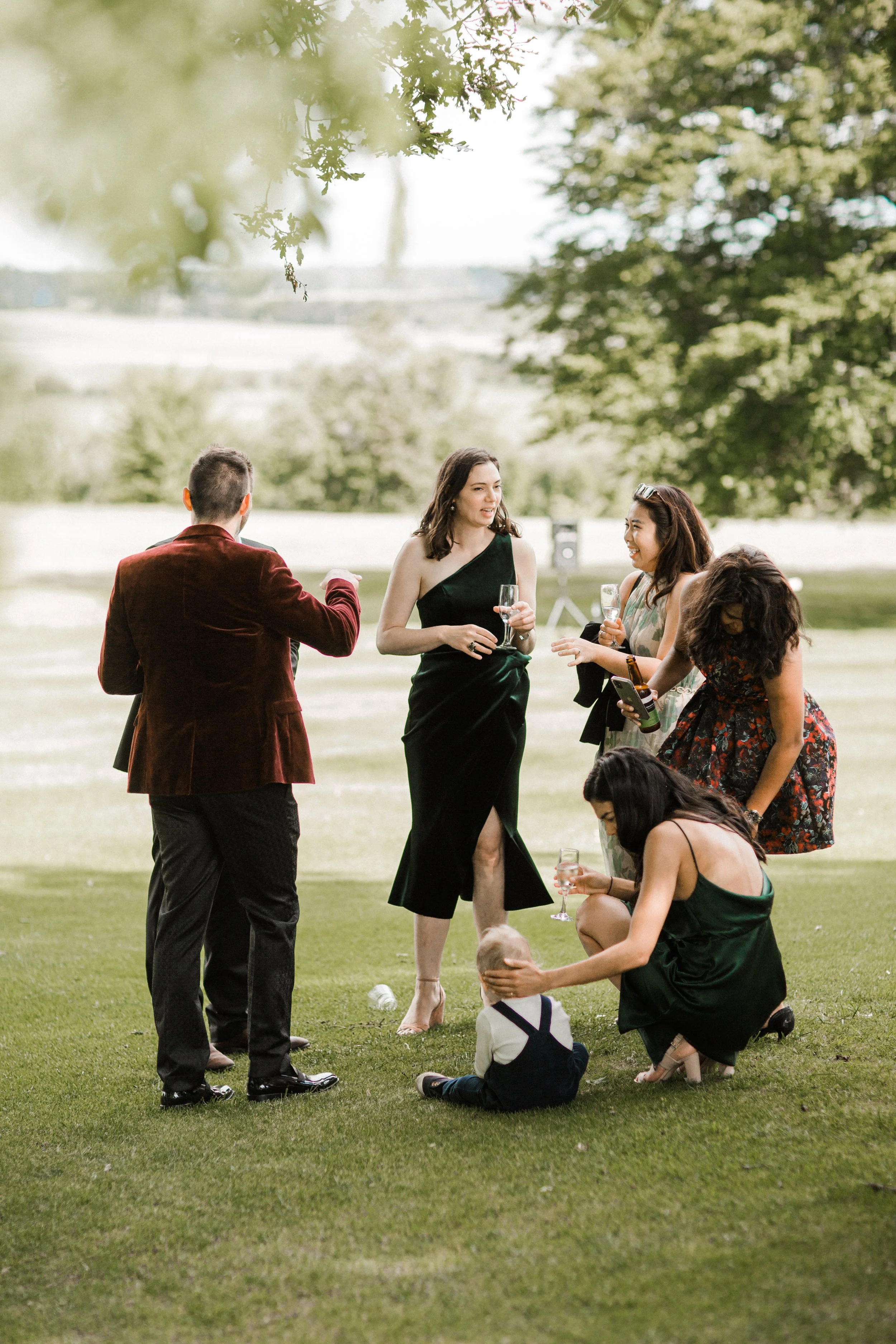 A group of six people, five women and one man, dressed in formal attire, are gathered outdoors on a grassy field with trees in the background. They are holding glasses of wine and engaging in conversation, with some sitting and some standing. One wom