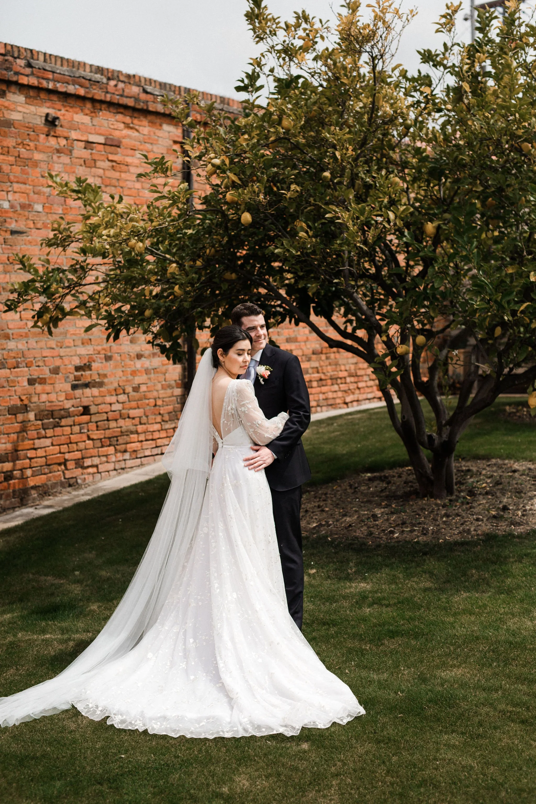 A bride and groom standing together outdoors, with the bride in a white wedding gown and veil, and the groom in a black suit and tie, next to a tree and a brick wall.