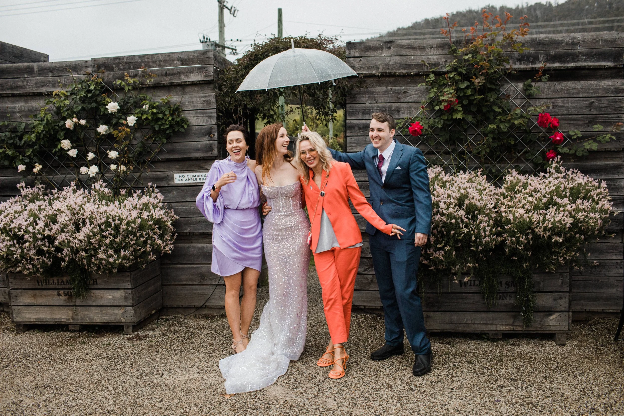 Four people celebrating outdoors under an umbrella, with a wooden fence and blooming bushes in background.