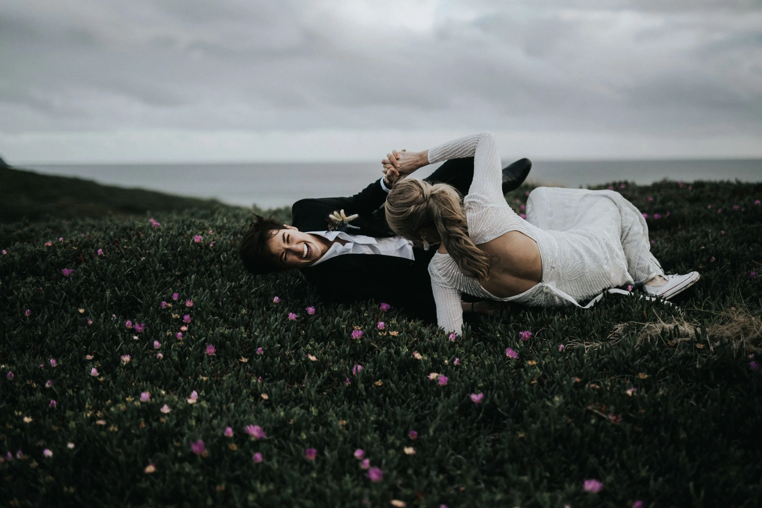 A couple dressed in wedding attire playfully wrestling on a grassy hill with pink flowers, overlooking the ocean under cloudy skies.