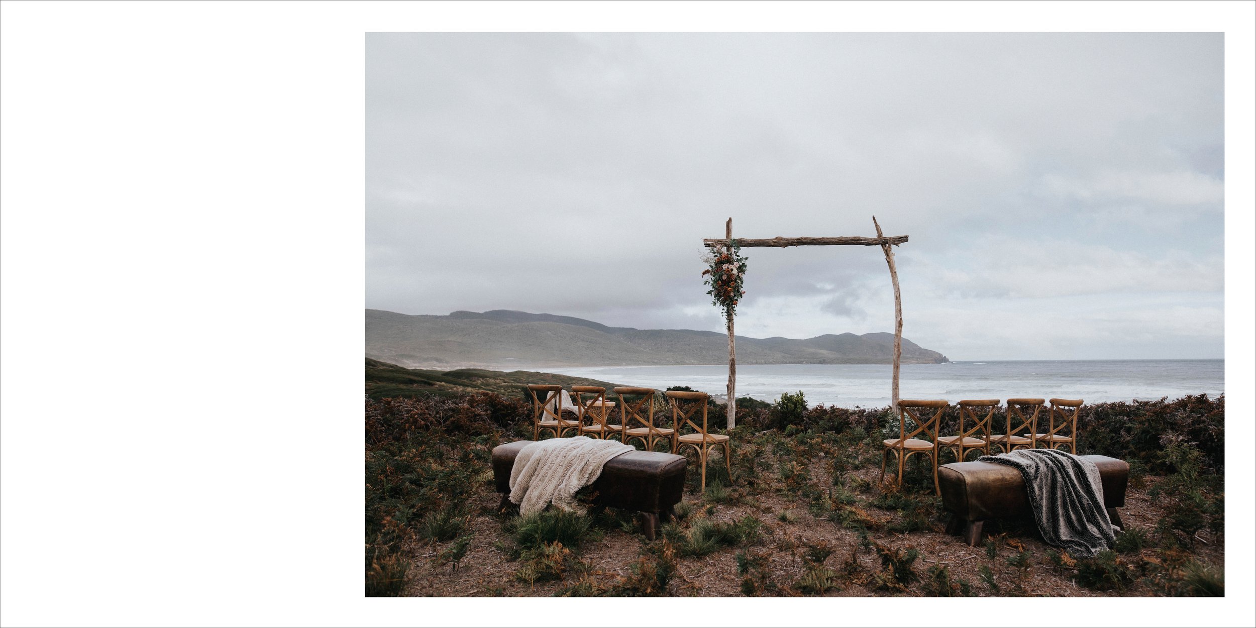 Outdoor wedding ceremony setup on a beach with wooden chairs and benches, a rustic wooden arch decorated with flowers, overlooking the ocean and distant hills under cloudy sky.