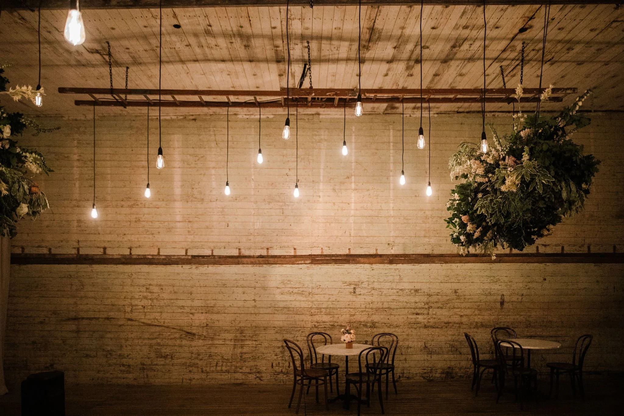 Interior of a rustic venue with hanging Edison bulbs, a large floral arrangement, and two small round tables with chairs.