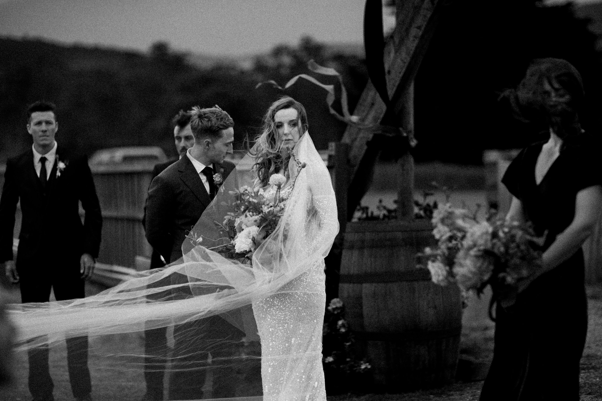 A black and white photo of a bride holding a bouquet walking outdoors during a wedding, with bridesmaids and groomsmen nearby.