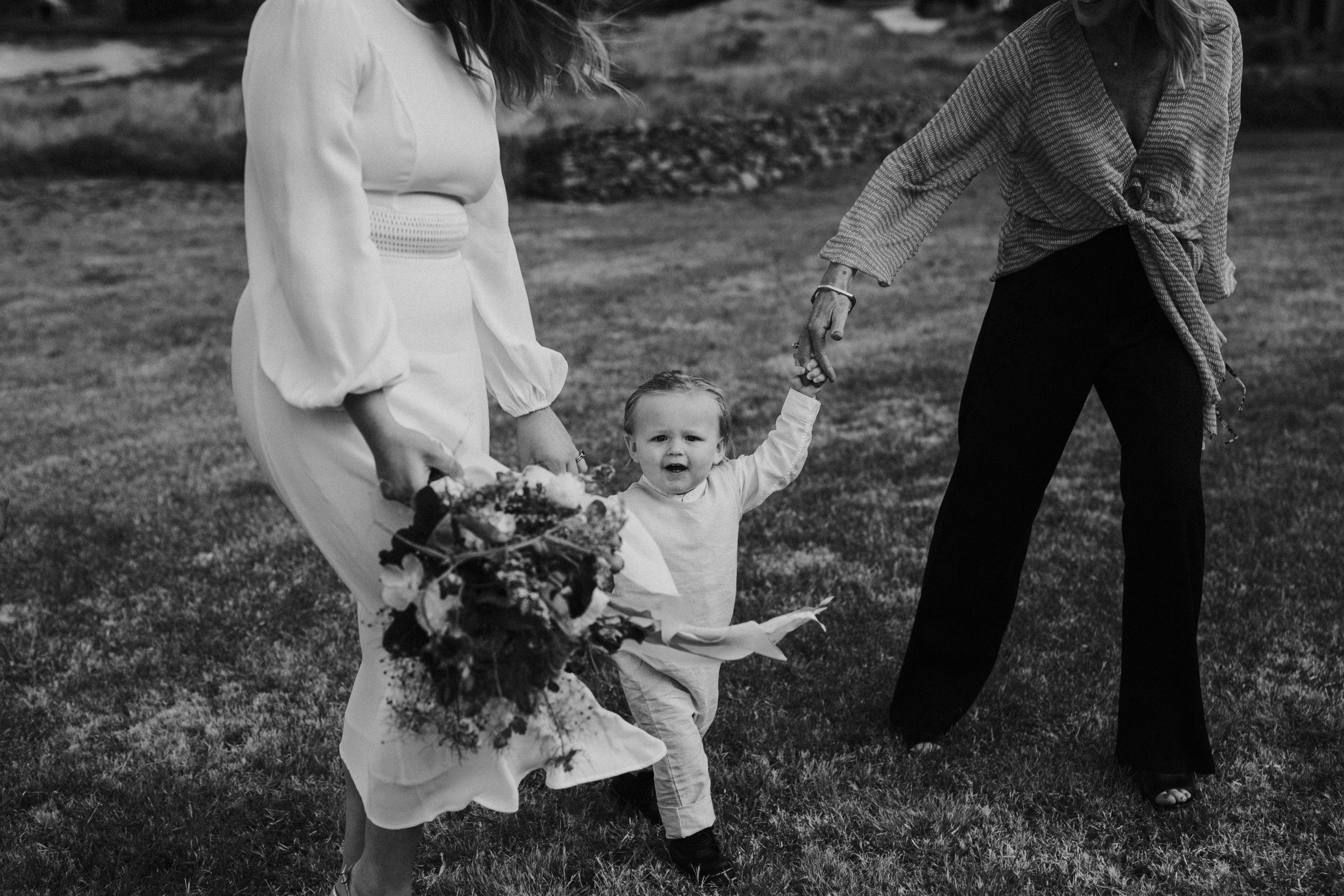 A young child walking between two women outdoors, holding their hands. One woman is holding a bouquet of flowers, and the scene appears to be a casual outdoor gathering.