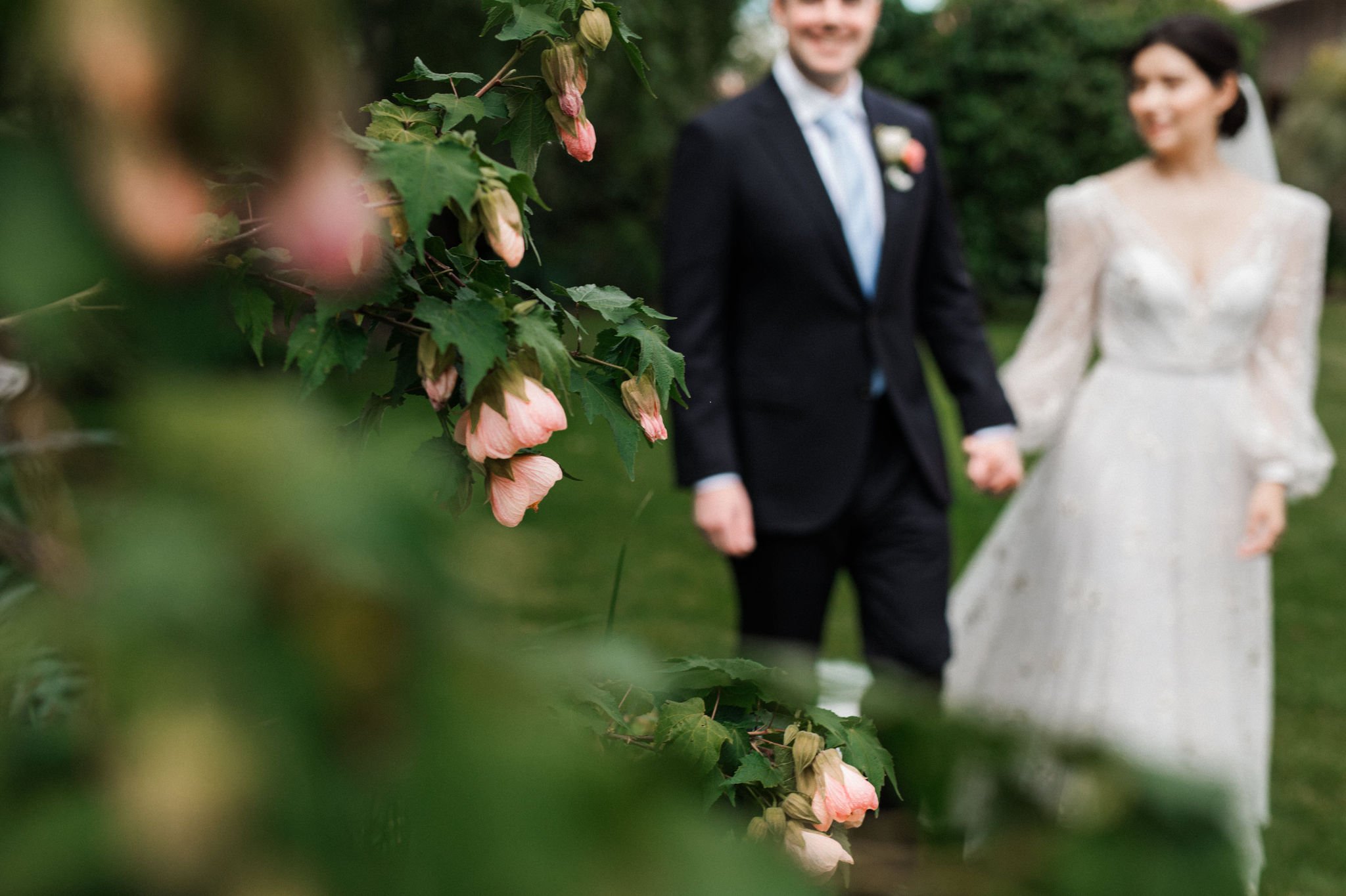 A bride and groom holding hands and walking outdoors, with pink flowers and green foliage in the foreground, slightly out of focus.