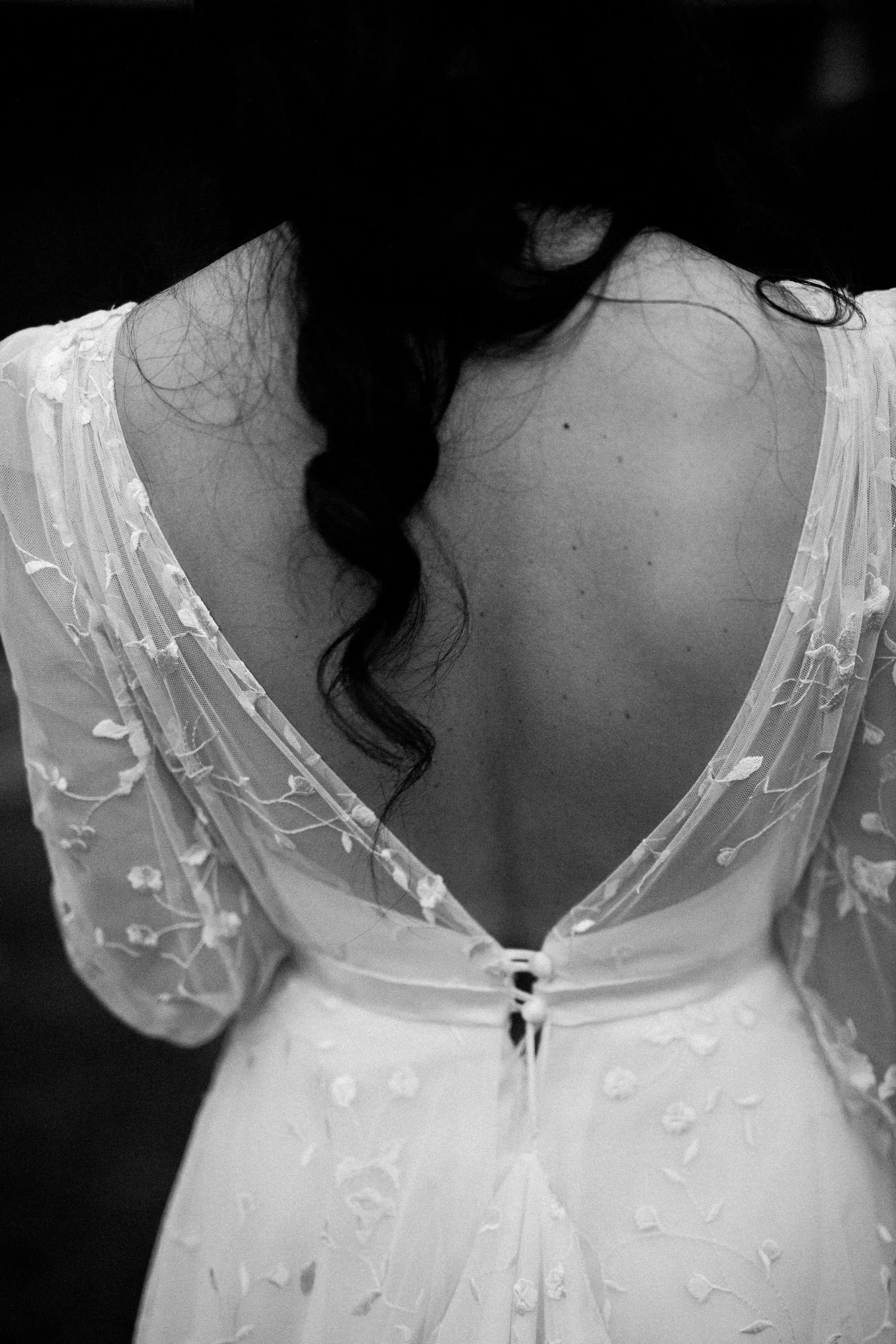 Close-up black-and-white photo of a woman wearing a sheer, embroidered dress with a deep neckline, facing away.