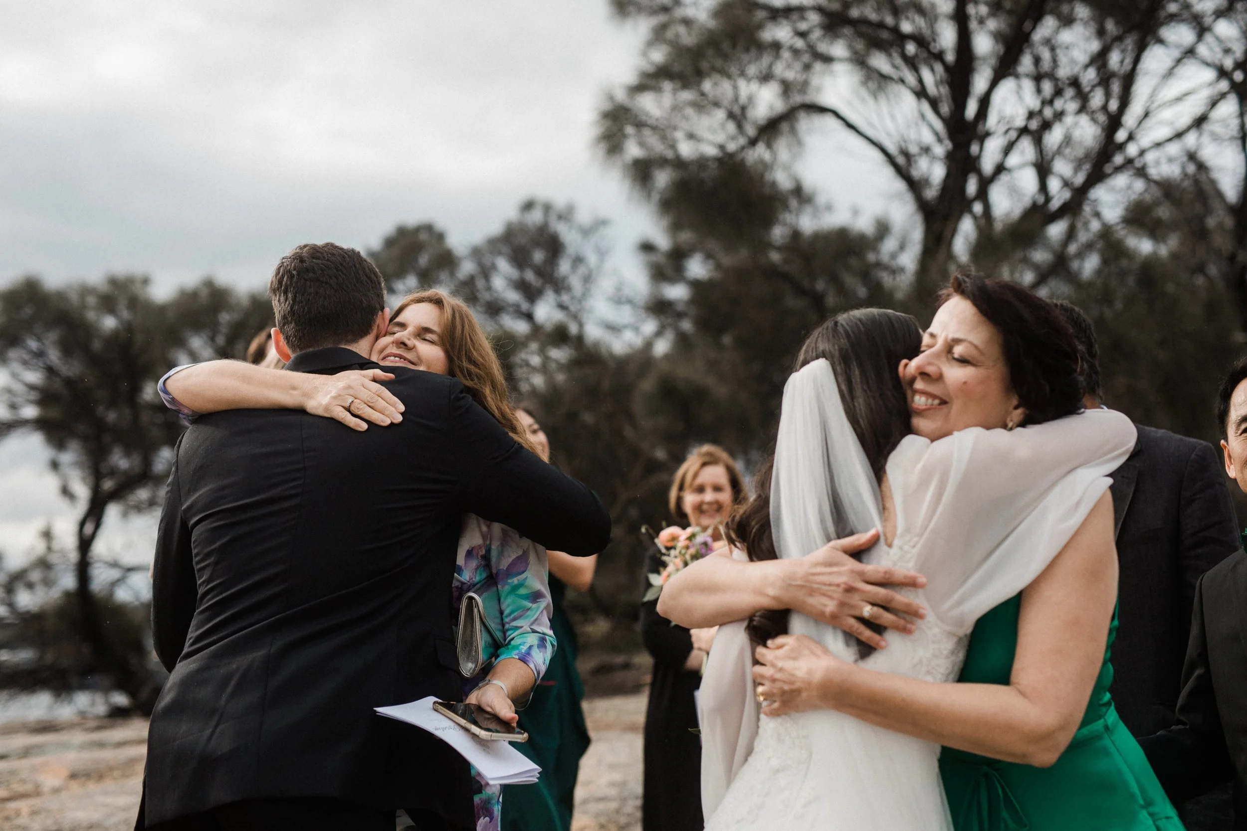 A group of people hugging outdoors during a wedding celebration, with trees and an overcast sky in the background.