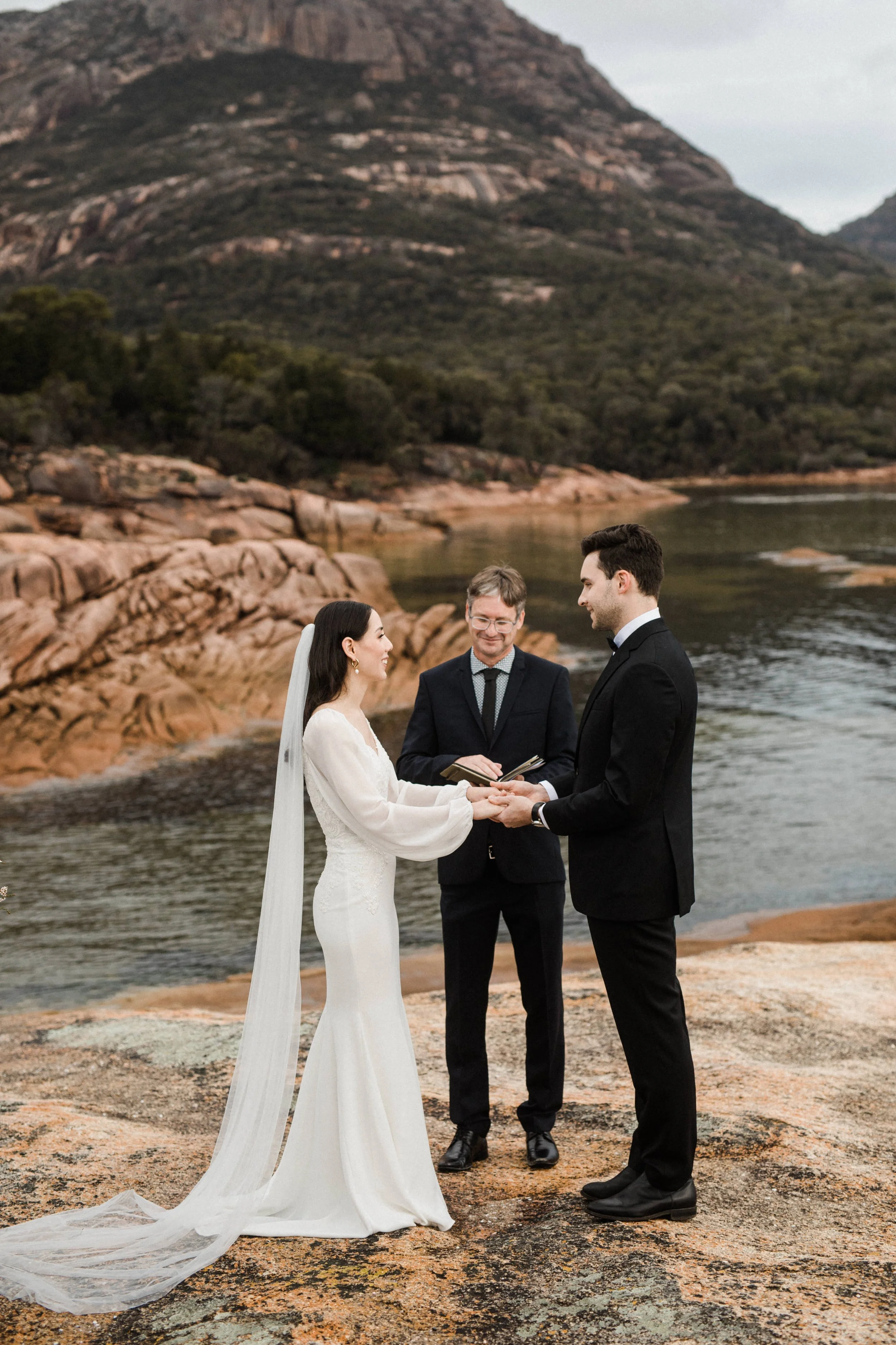 A wedding ceremony taking place outdoors on a rocky shoreline with mountains in the background. The bride and groom are holding hands and exchanging vows, with an officiant present.