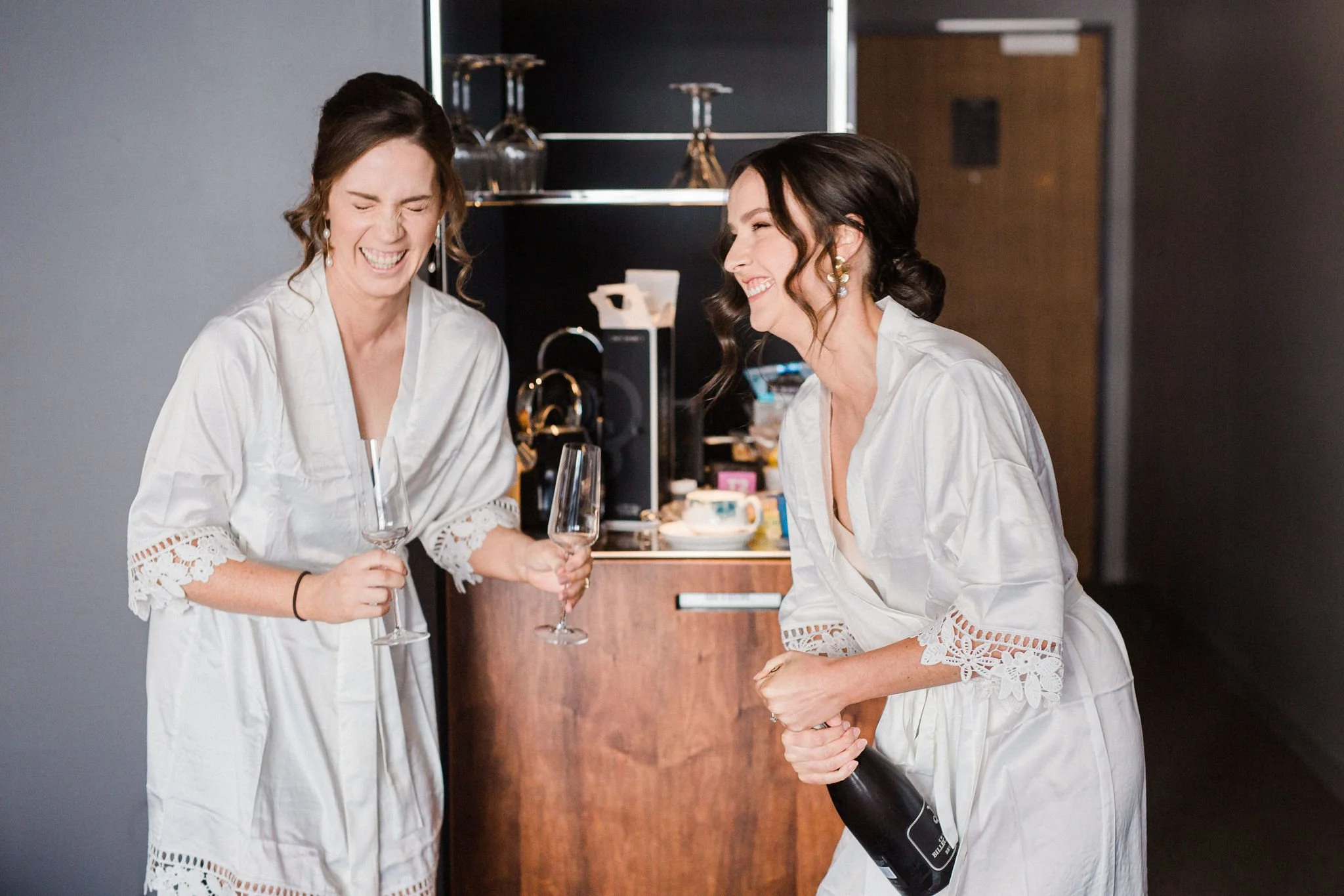 Two women in white robes laughing and holding champagne glasses, one woman also holding a bottle of champagne, in a room with a dark wall and a wooden cabinet.