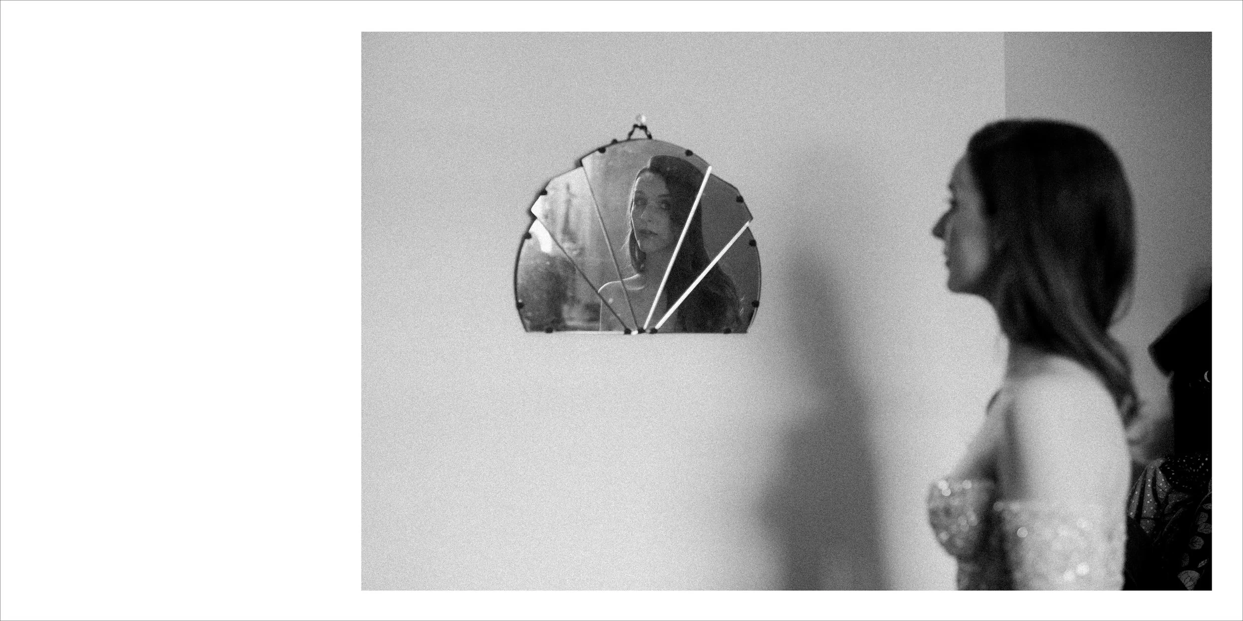 A woman in an off-shoulder dress is looking at her reflection in a decorative mirror on a plain wall.