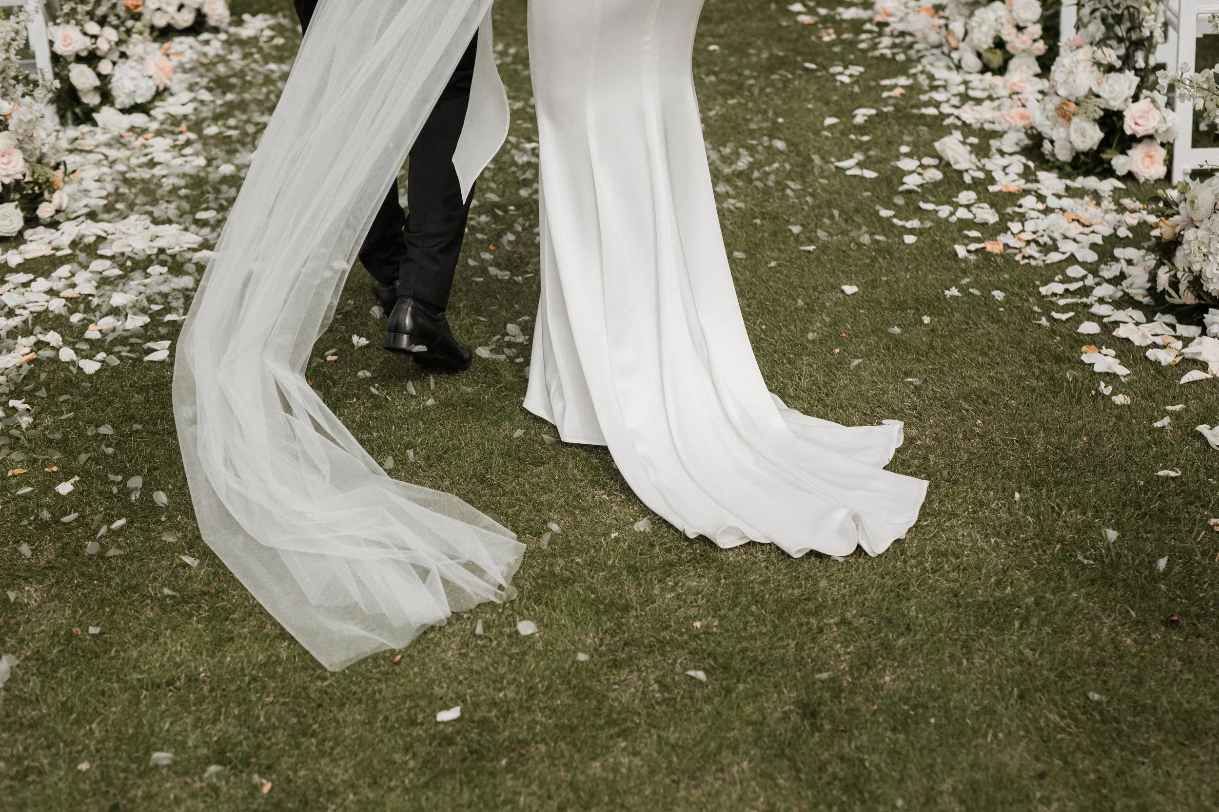 Close-up of a bride and groom walking on grass after their wedding ceremony, with flower petals scattered on the ground and floral arrangements along the aisle.