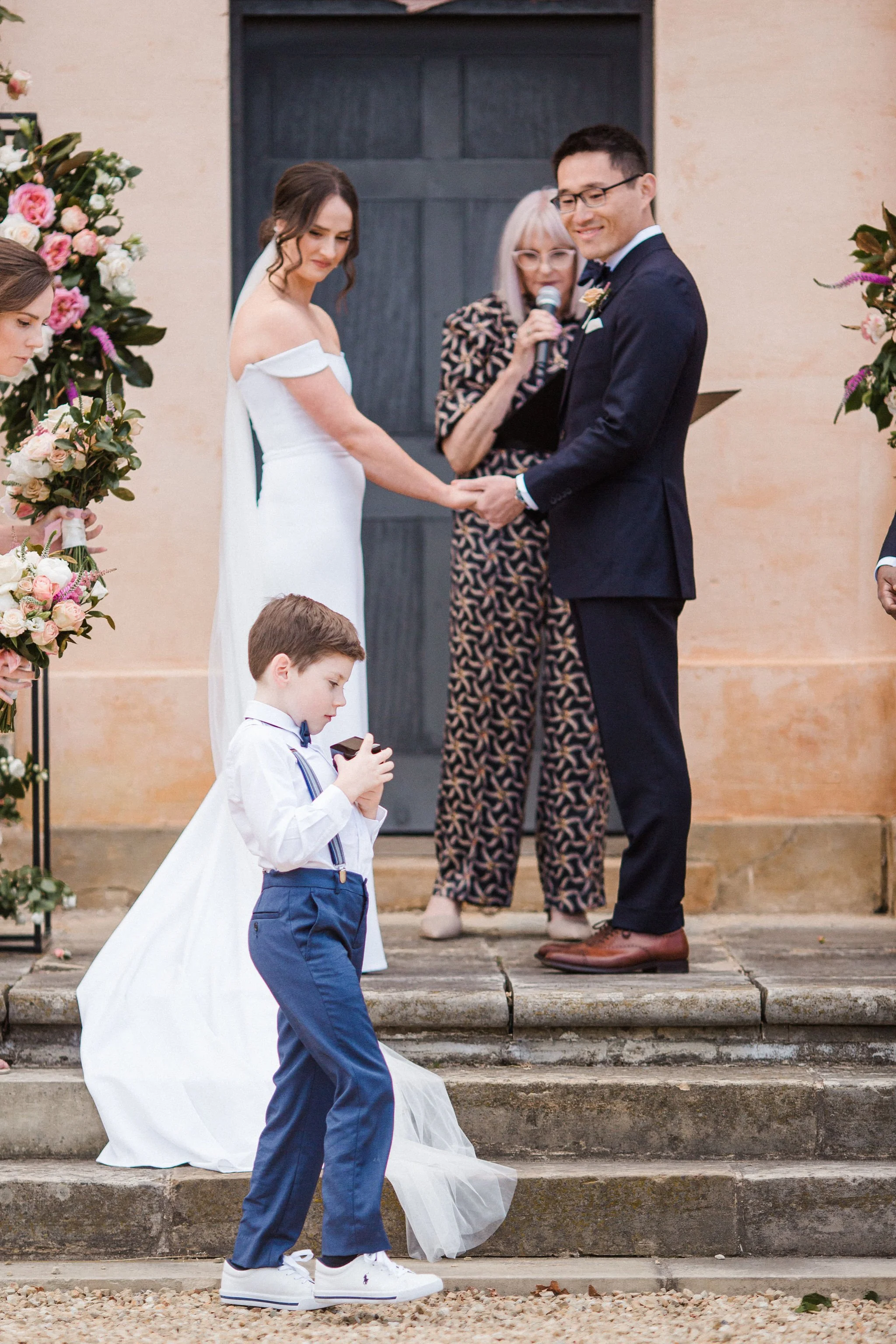 A bride and groom holding hands during their wedding ceremony with an officiant behind them. A young boy in white shirt and blue pants stands in the foreground, looking at a device.