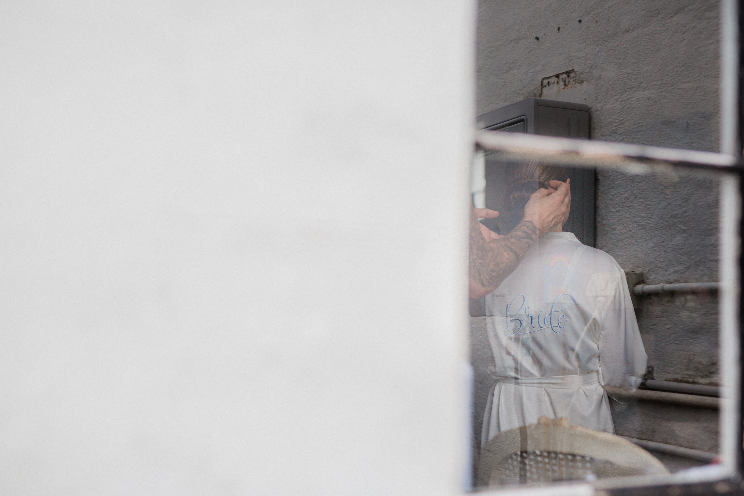 A woman in a white bathrobe with more tattoos on her arm is looking in a mirror held by a person whose face is not visible, with the word 'Bride' embroidered on the back of her robe.