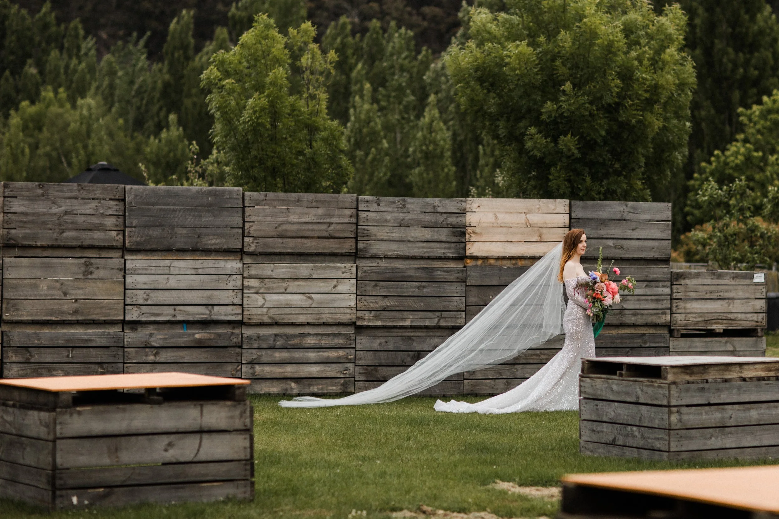 Bride in a white wedding gown with a long veil holding a bouquet of flowers standing outdoors near a wooden fence.