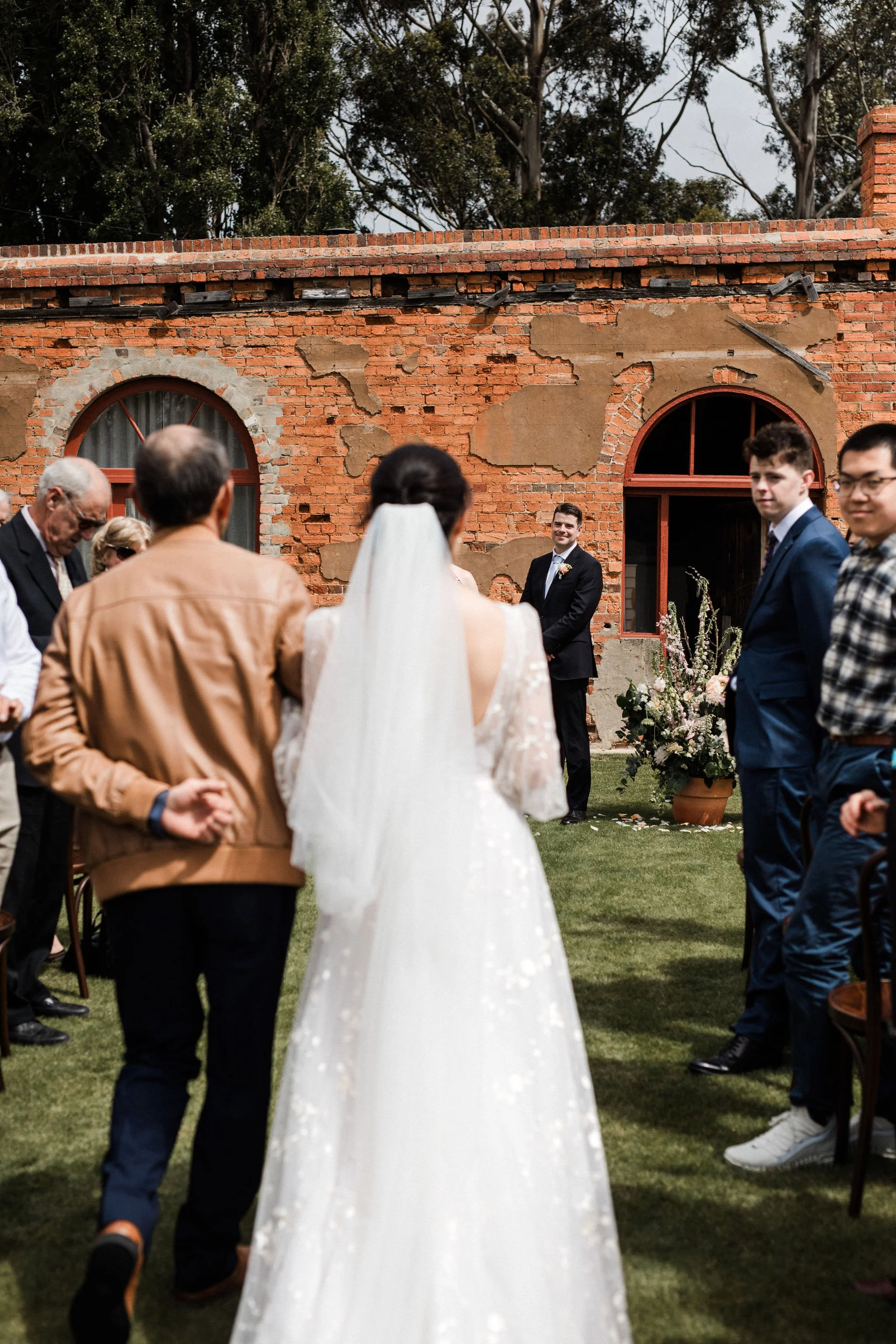 A bride with a white veil and dress walking down the aisle towards a groom in a black suit and tie during an outdoor wedding ceremony in front of a weathered red brick wall and lush trees.