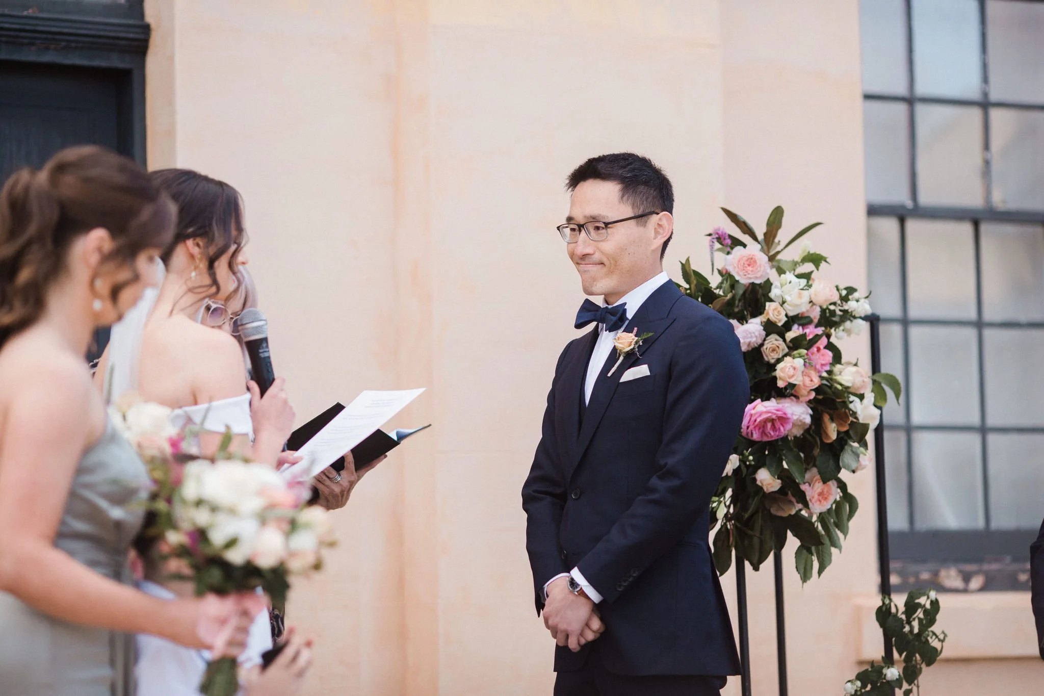 A groom in a black tuxedo with a bow tie and glasses standing at a wedding ceremony, facing a woman holding a microphone and reading from a paper. The woman and others in the foreground are out of focus, holding bouquets of flowers, with a large flor