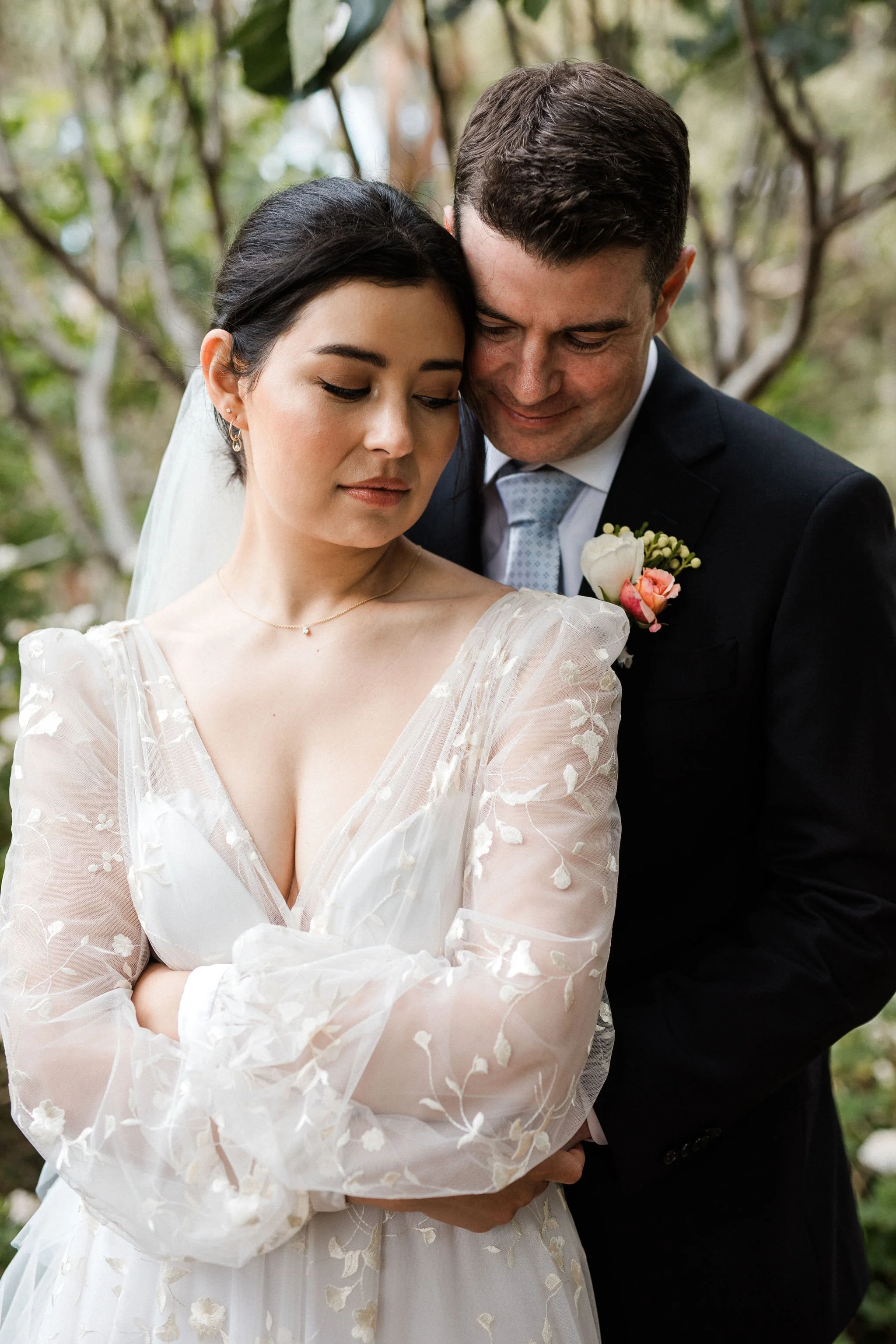 A bride and groom embrace outdoors, with the bride in an embroidered white wedding dress and the groom in a dark suit with a boutonnière, surrounded by trees.