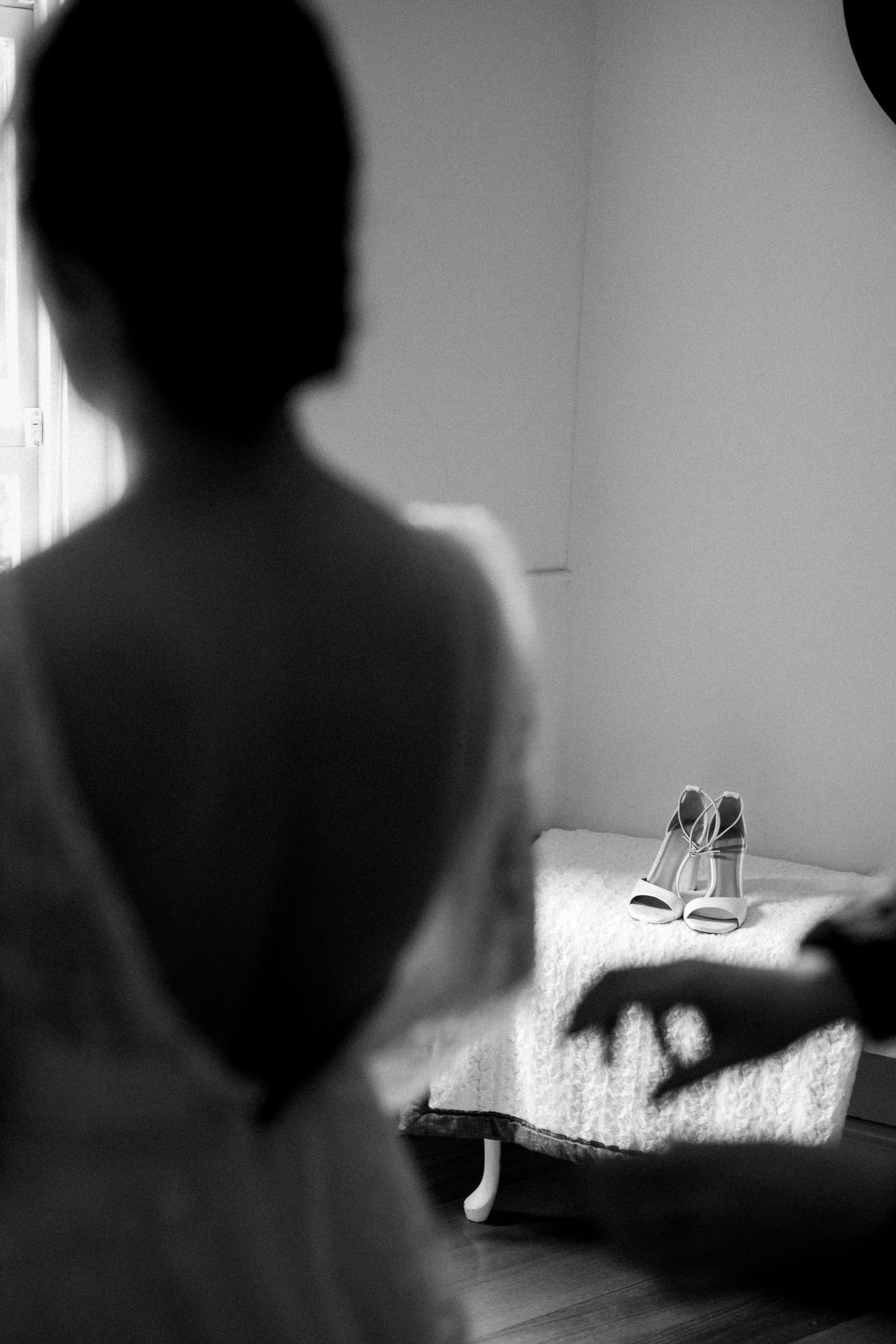 Black-and-white photo of a person with short hair in the foreground, facing away, with a pair of high-heeled shoes on a bed in the background.