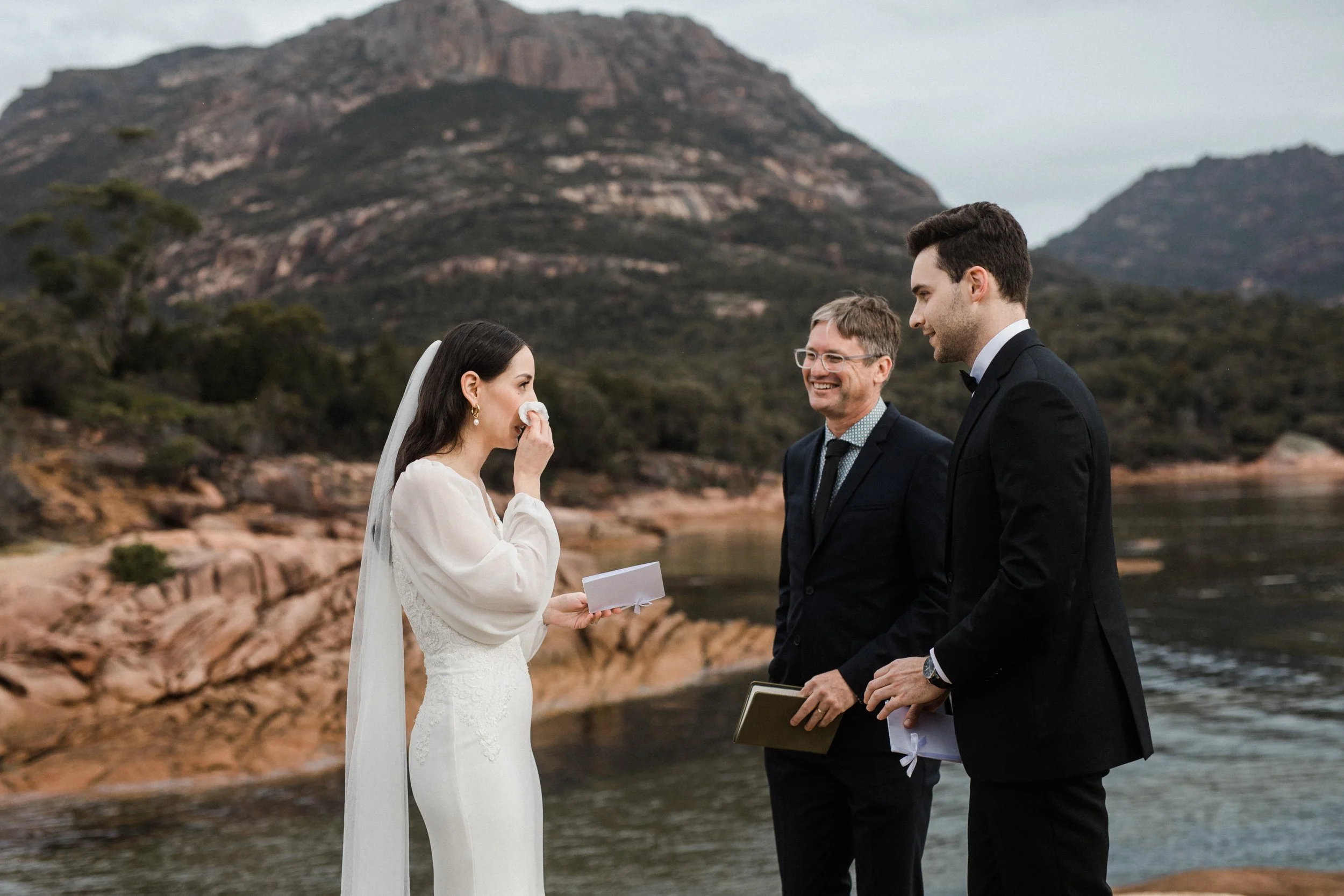 A woman in a wedding dress crying during a wedding ceremony by a river with mountains in the background, as a man and officiant look on.