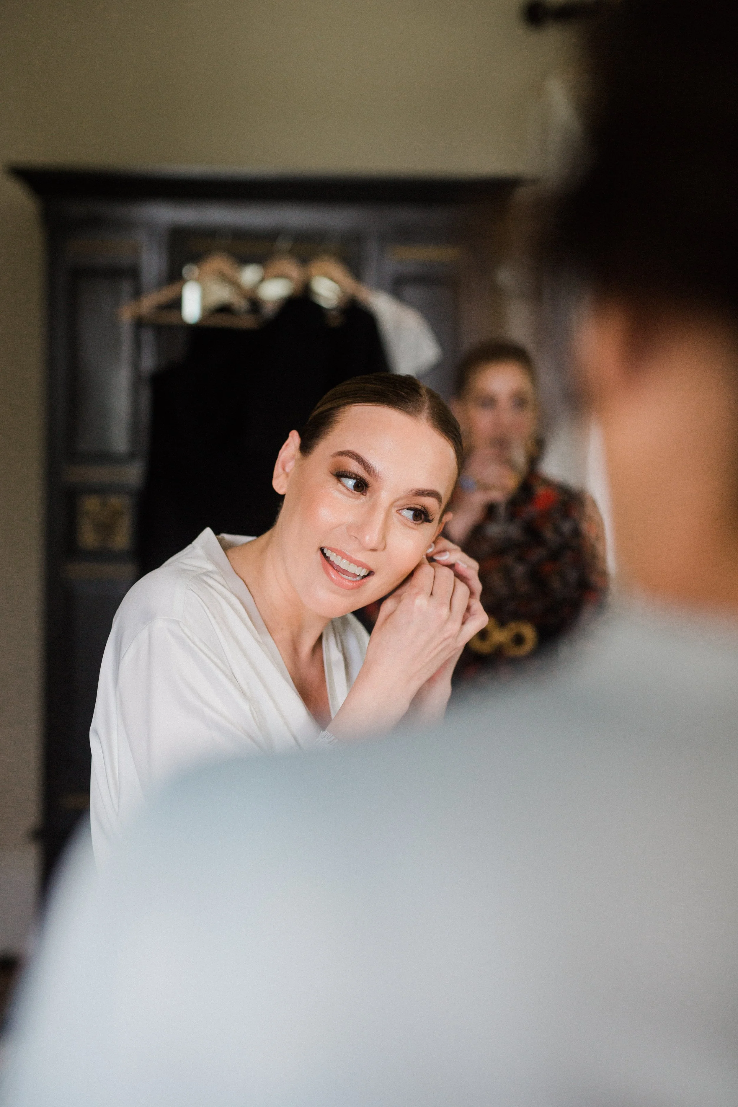 A woman with makeup sitting in front of a mirror, smiling as she adjusts her earring, with another woman in the background holding a glass.