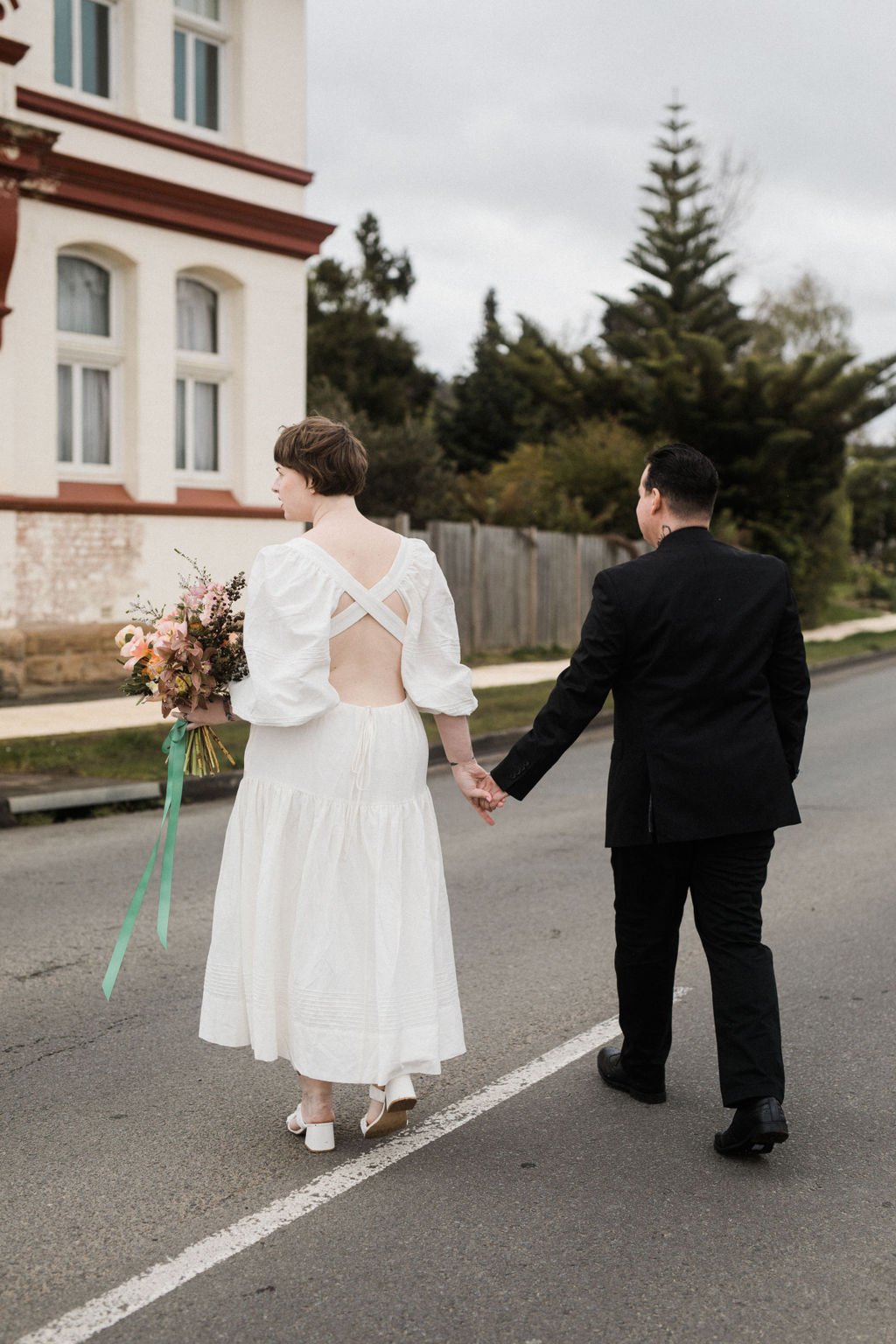 A bride and groom holding hands and walking on a street. The bride is wearing a white gown with an open back and is holding a bouquet. The groom is dressed in a black suit. They are walking away from the camera, towards a residential area with trees 