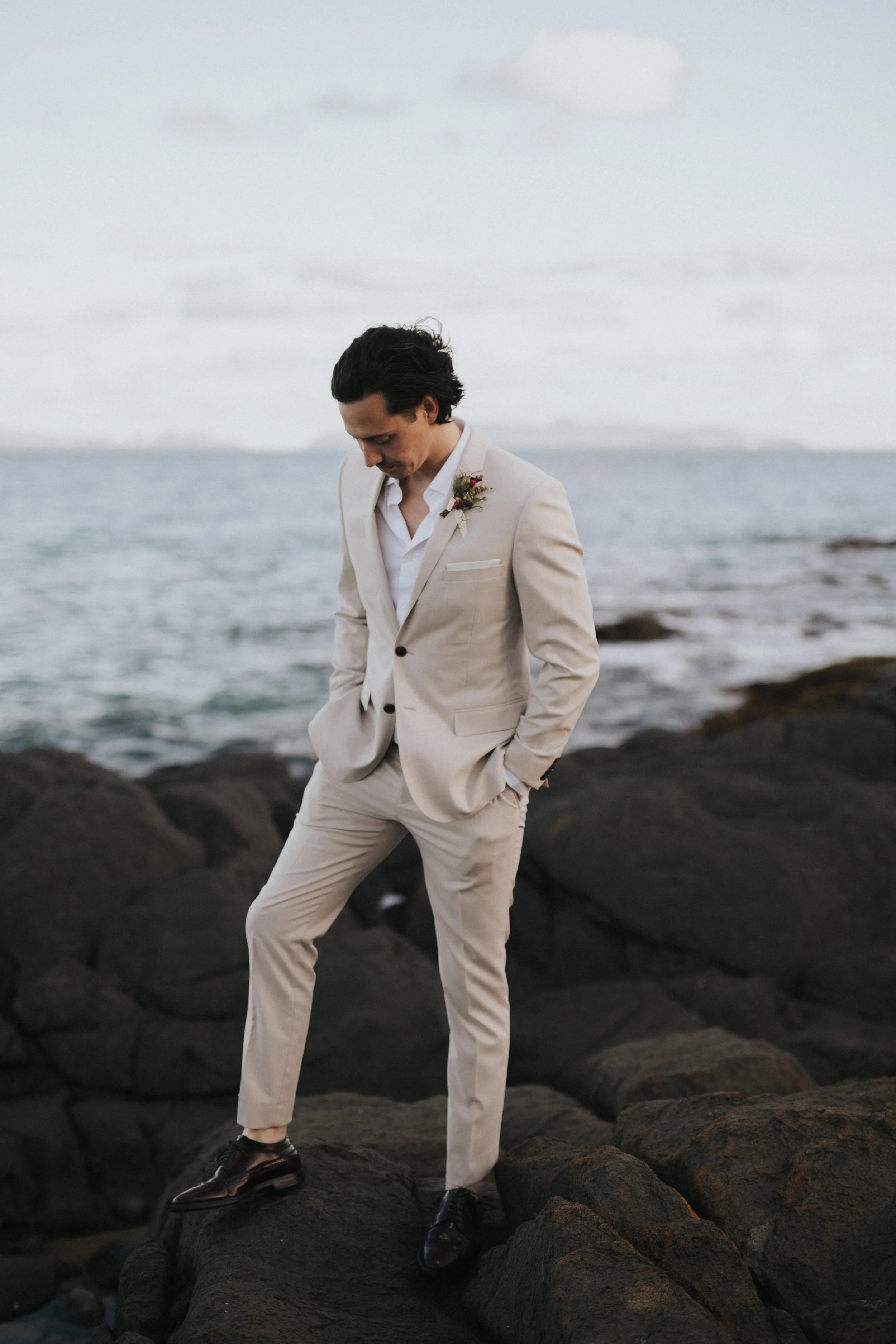 A man dressed in a light beige suit with a boutonnière, standing on rocks by the ocean with his hands in his pockets, looking down.