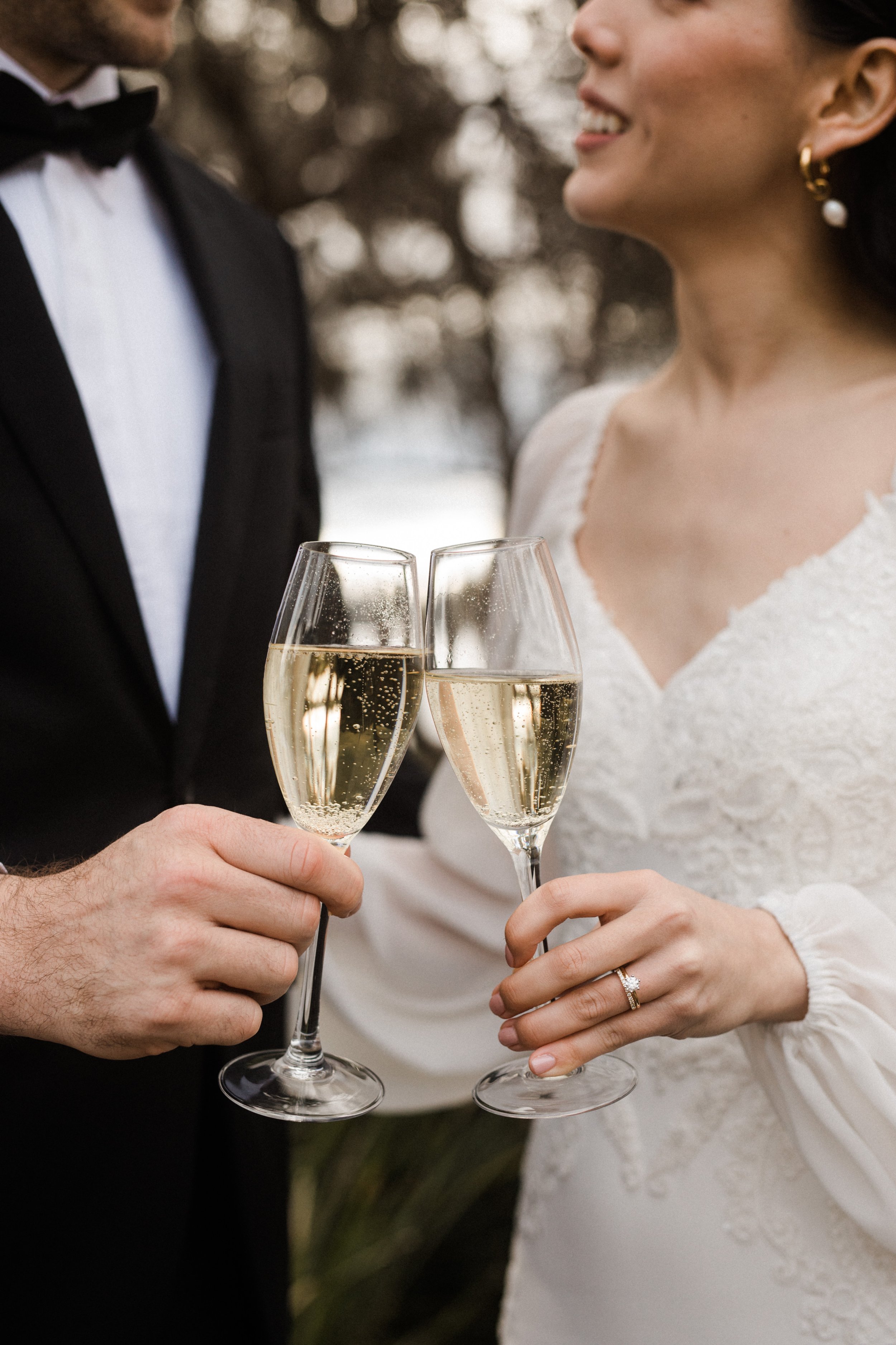 A close-up of a bride and groom holding champagne glasses for a toast at their wedding, with the bride wearing a white dress and a wedding ring, and the groom in a black tuxedo with a bow tie.